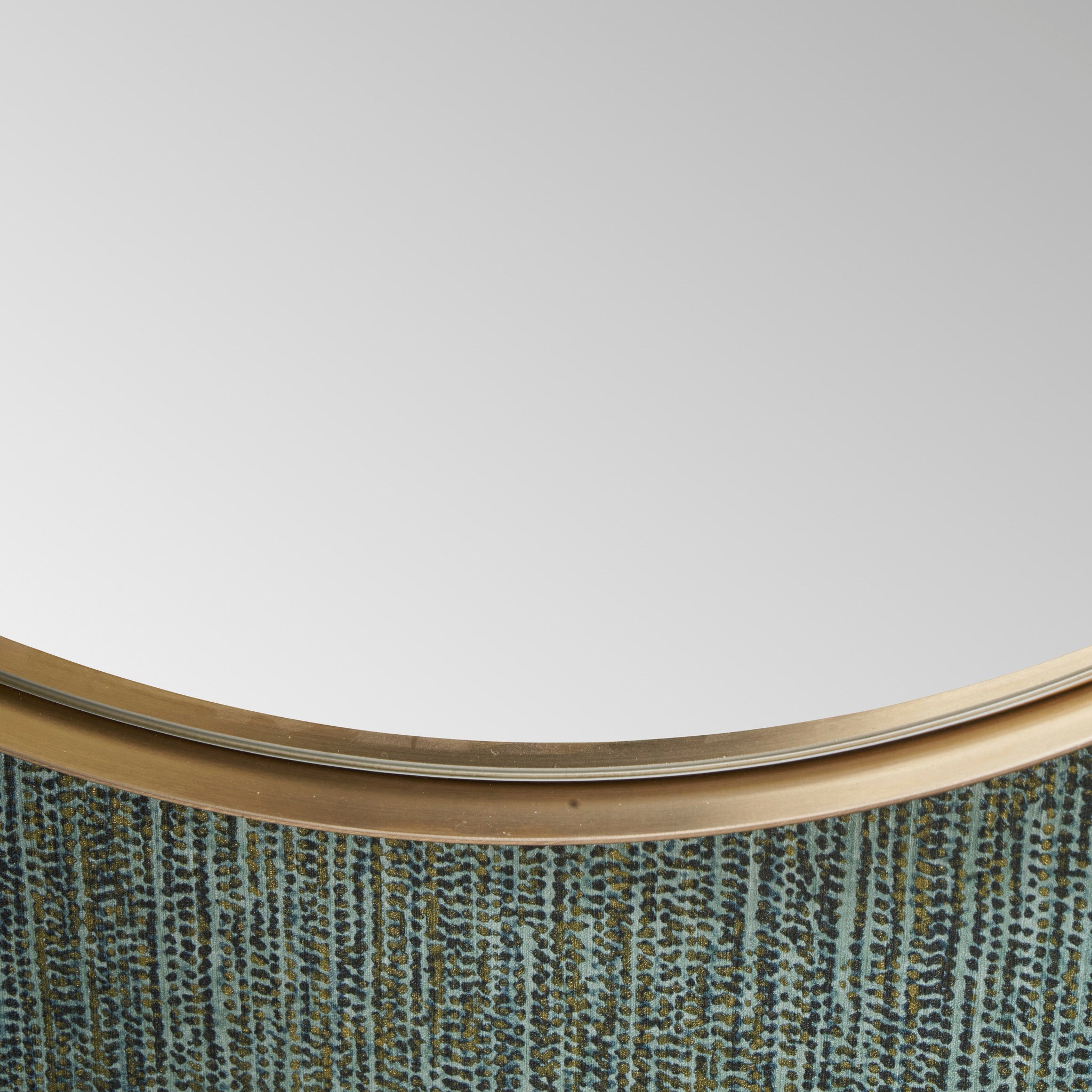 Close-up of the Brushed Gold Metal Slim Frame Round Wall Mirror Medium, reflecting a plain white surface, mounted on a textured background.