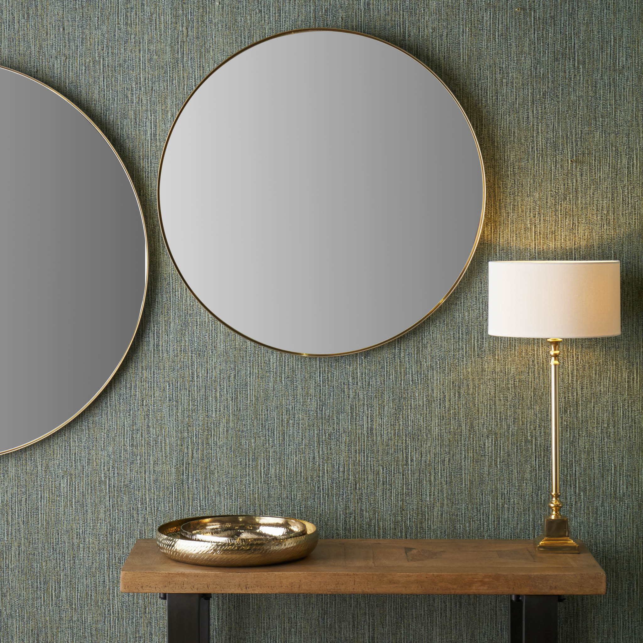 The Brushed Gold Metal Slim Frame Round Wall Mirror Medium pairs beautifully with a tall lamp and decorative bowl on a wooden table set against the textured green wall.