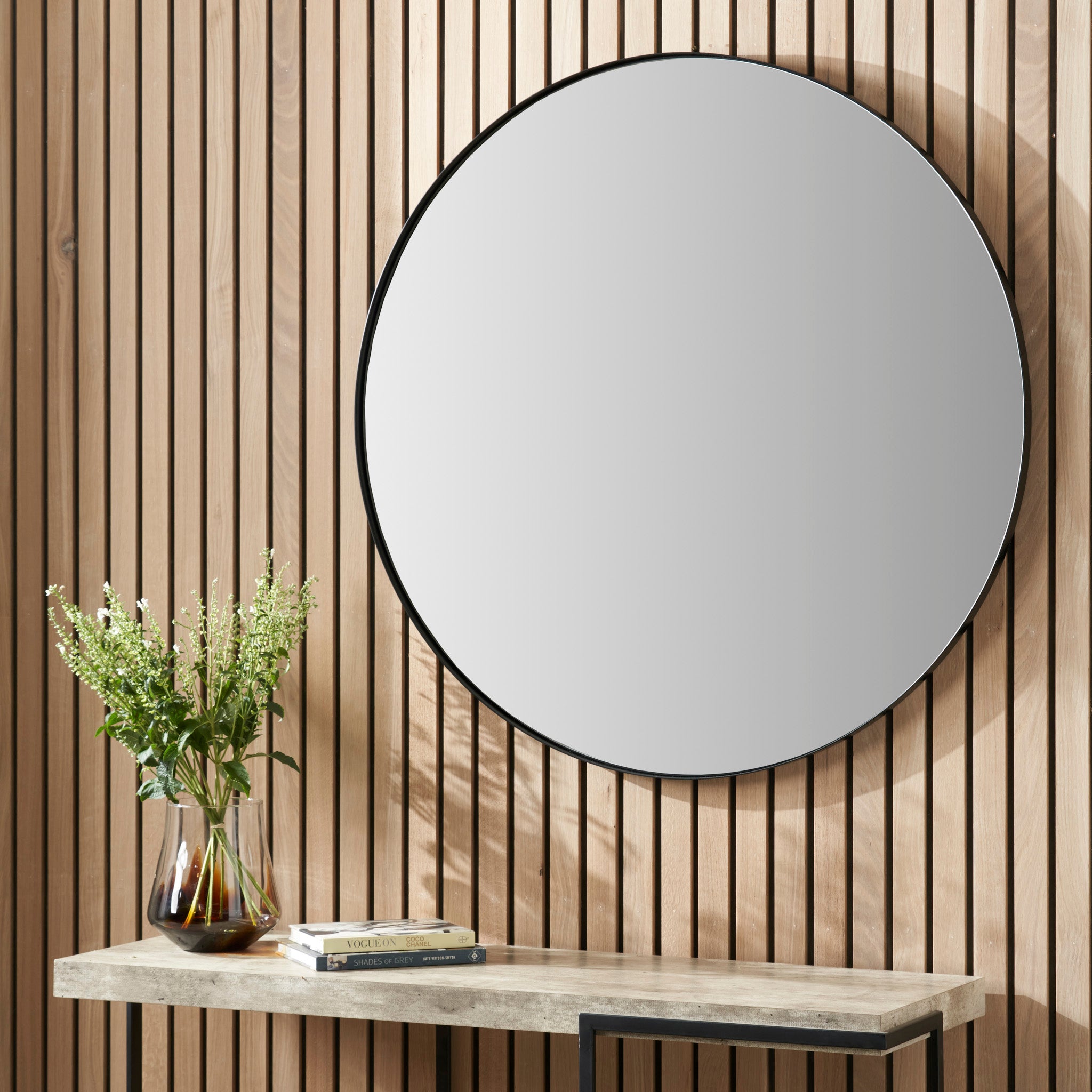 A Black Metal Slim Frame Round Wall Mirror Large is displayed on the wooden slat wall above a console table decorated with a vase of flowers and books.