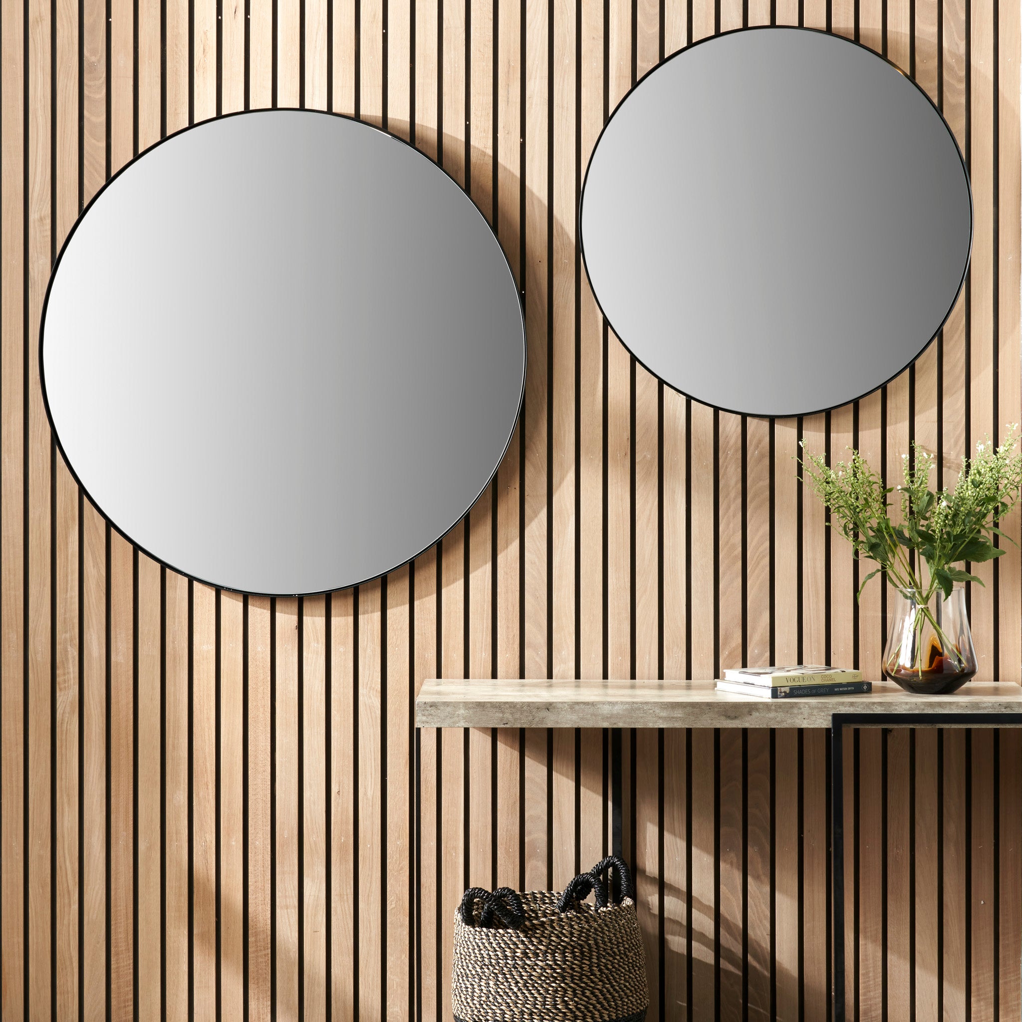 A pair of large black metal slim frame round wall mirrors decorate a wooden slat wall. Below them, a concrete console table is paired with a basket and a vase filled with flowers, creating an elegant display.