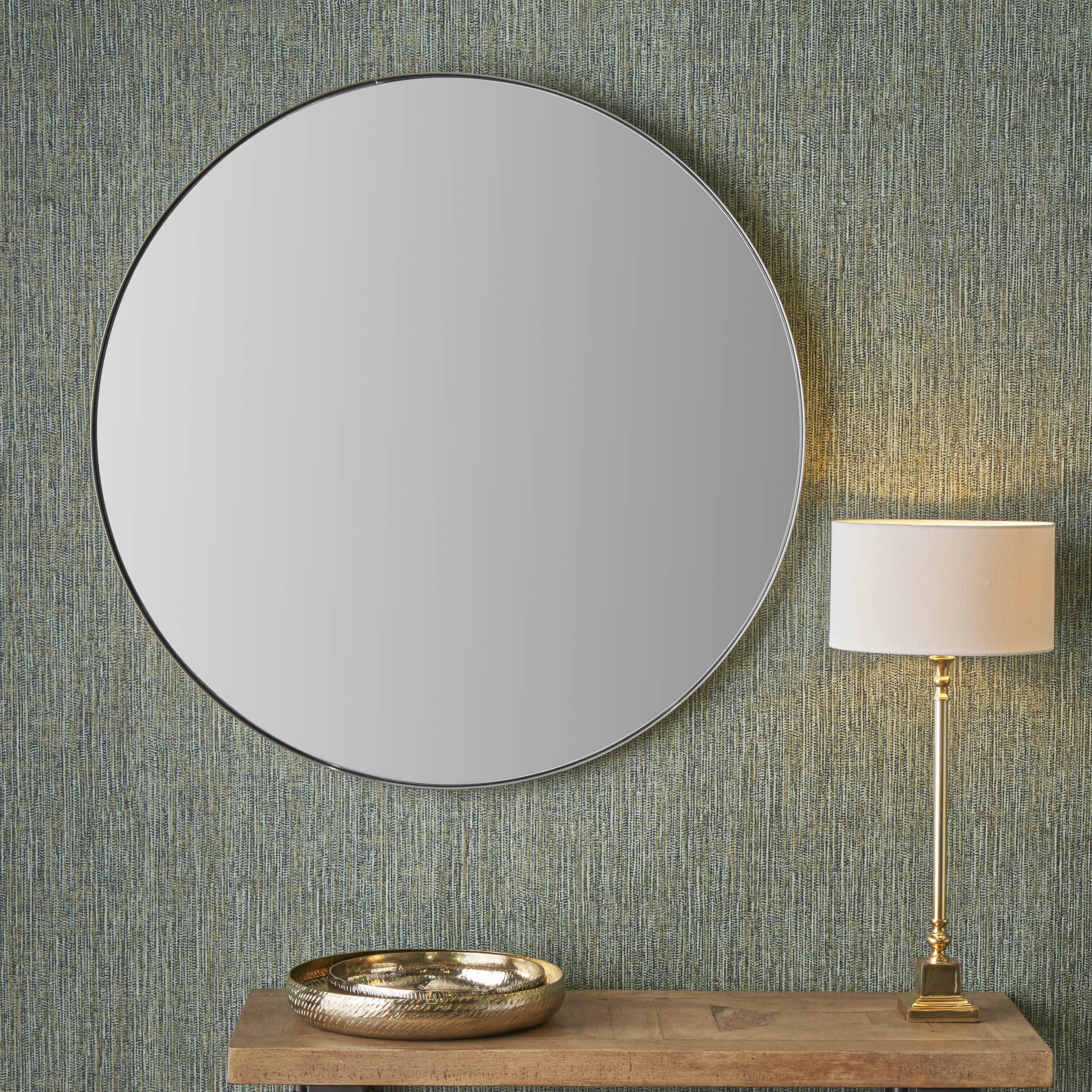 A large brushed silver round wall mirror with a slim frame hangs on the textured wall above a wooden table adorned with a gold tray and a tall lamp, creating an elegant contrast in matt black.