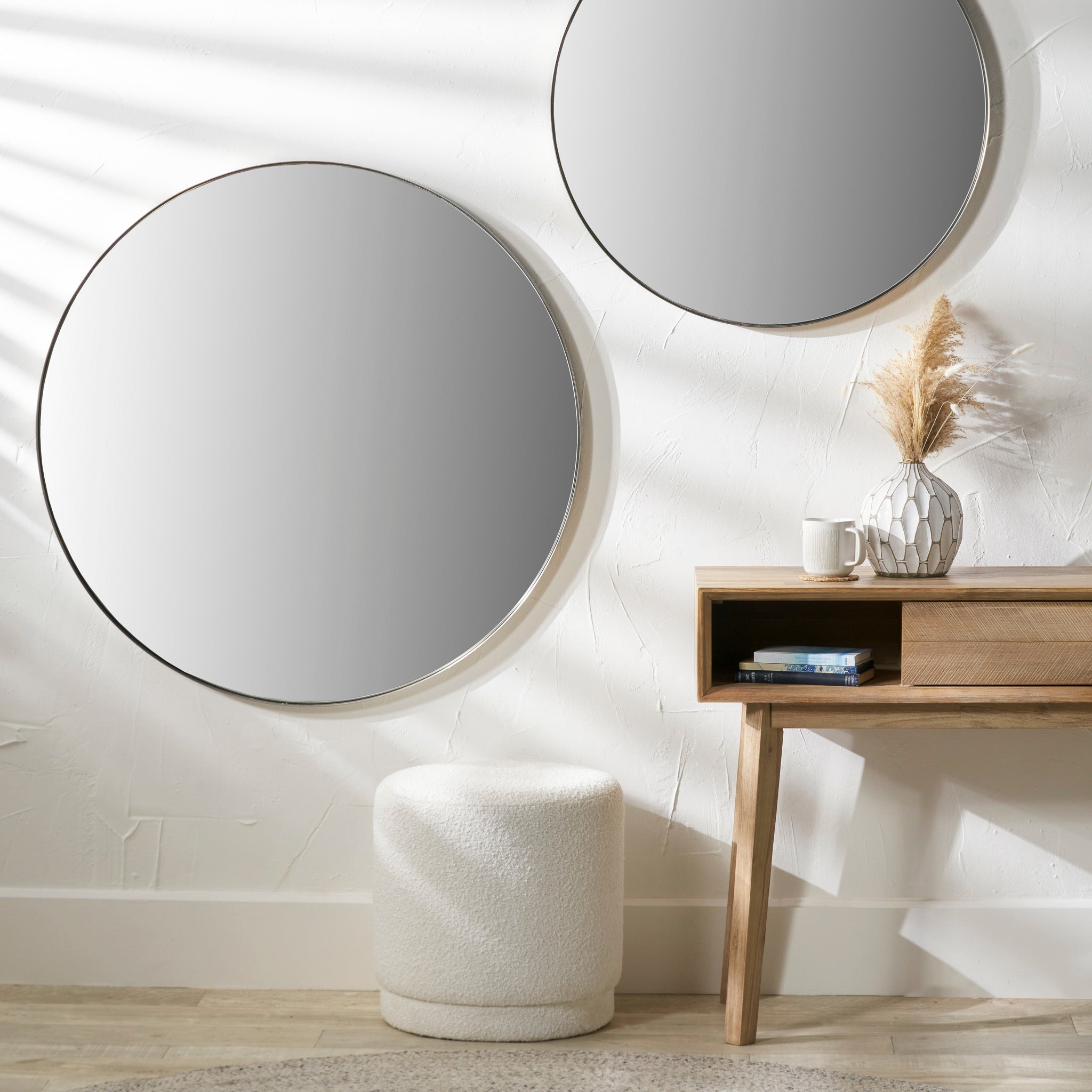 A large Brushed Silver Metal Slim Frame Round Wall Mirror adorns a textured wall above a wooden table, which holds a vase, cup, and books; a soft stool sits nearby. The matte black finish adds elegance to the space.