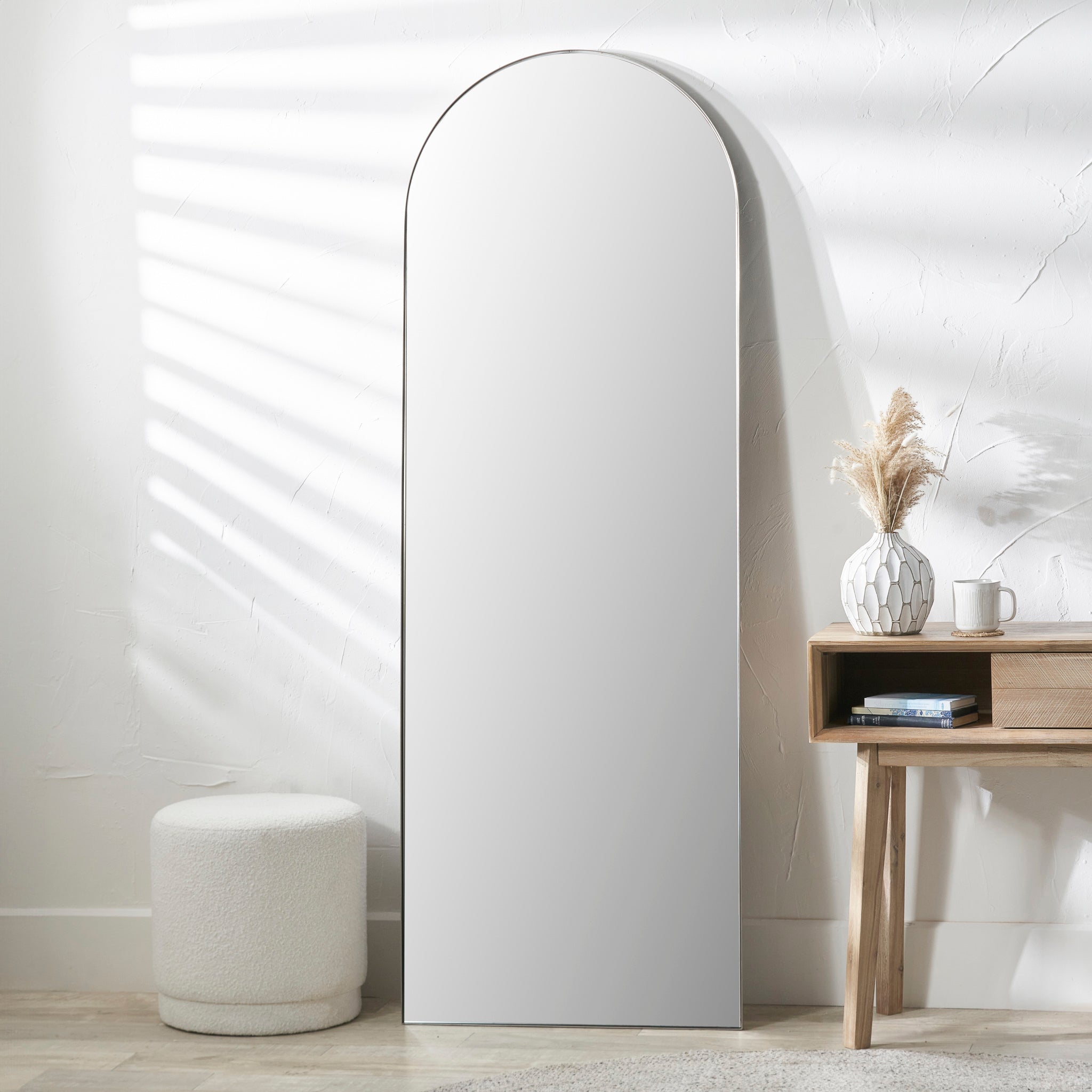 The Pacific Lifestyle Brushed Silver Metal Slim Frame Arch Floor Mirror stands by the wooden table with a vase and books. Soft light streams through blinds, illuminating the round stool on the floor—a perfect blend of minimalist decor.