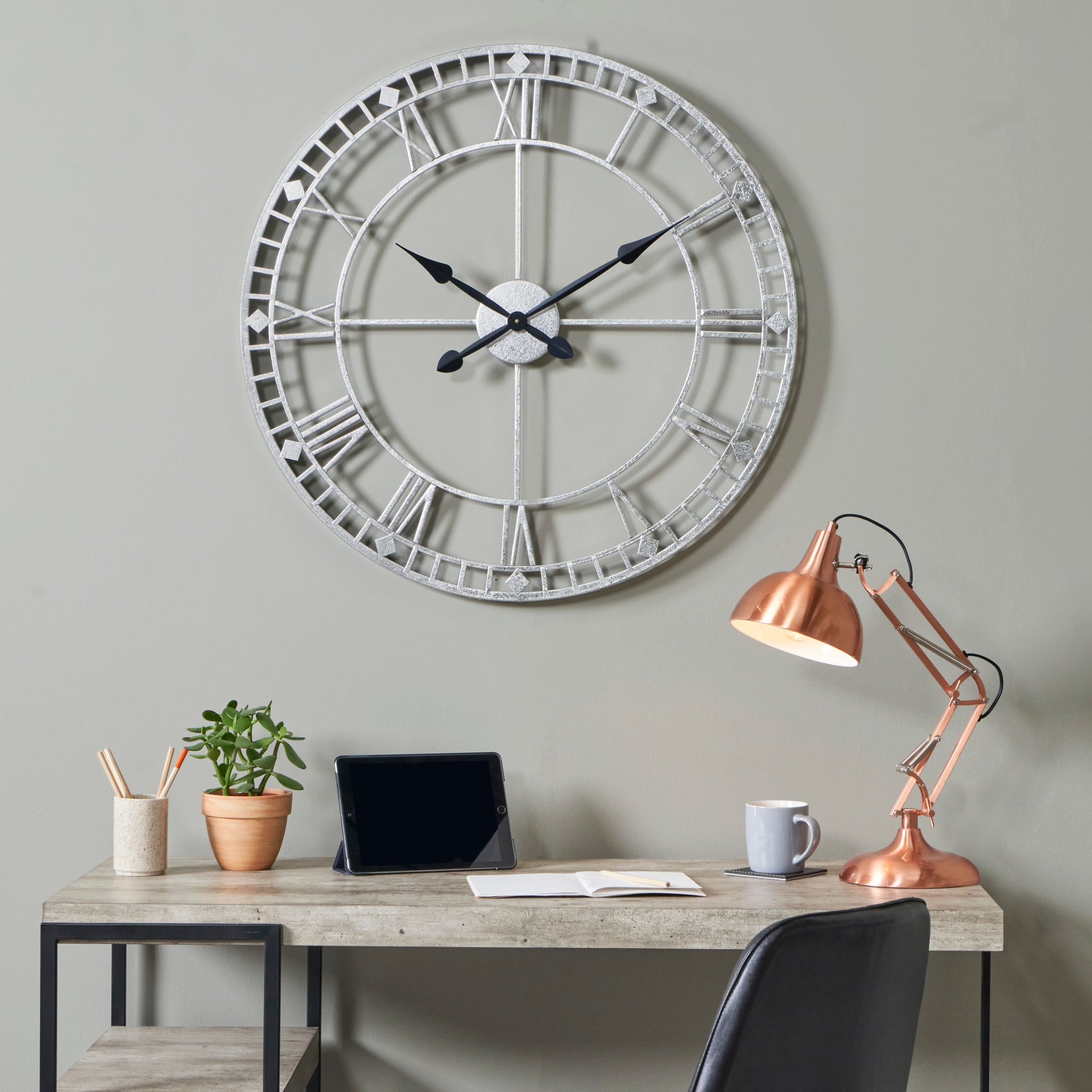 A desk holding a plant, notebook, lamp, and tablet sits beneath the Antique Silver Metal Round Wall Clock Large on a gray wall. The clock's Roman numerals infuse the modern workspace with a classic touch.