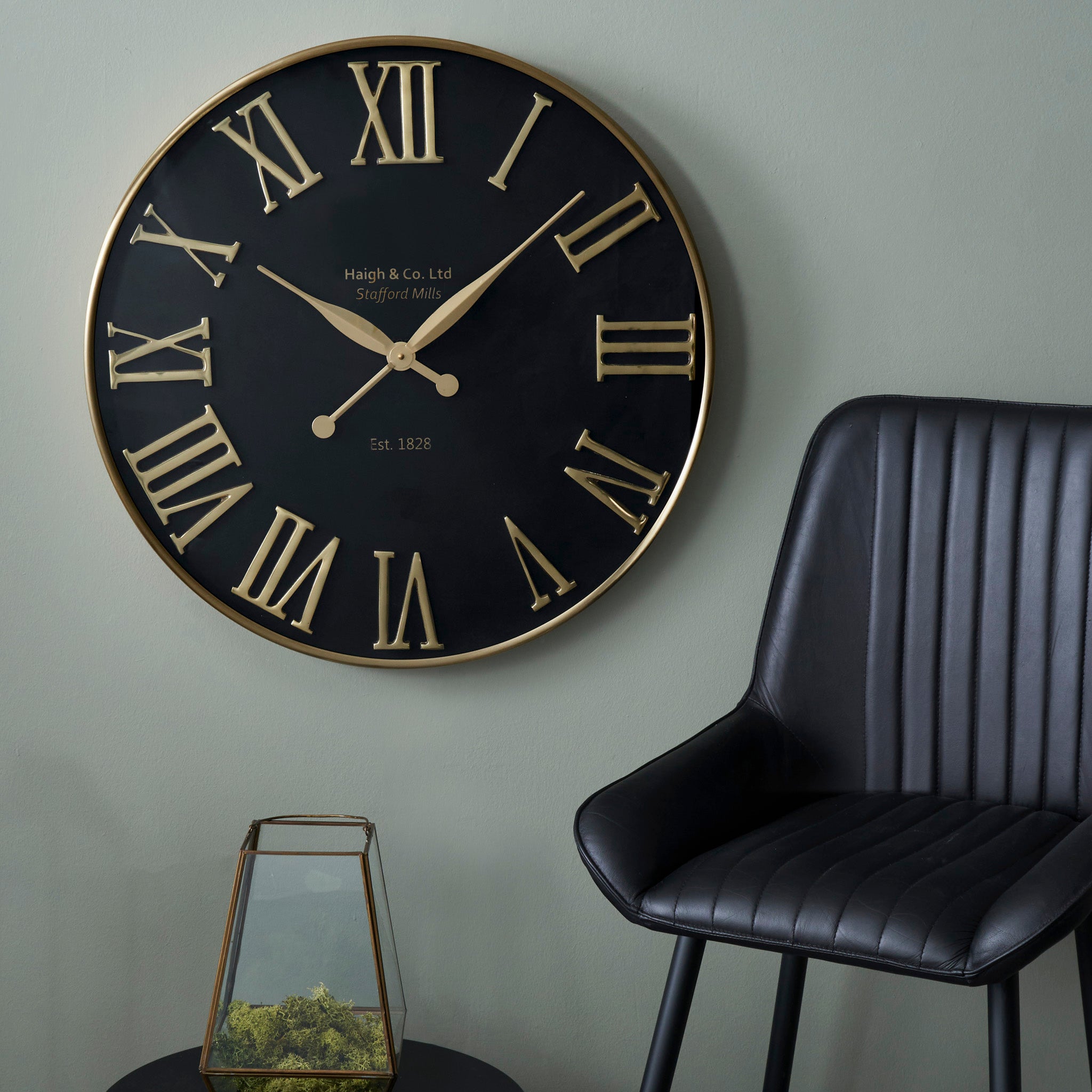 The Antique Gold and Black Metal Wall Clock with oversized Roman numerals graces the wall, perfectly complementing the black chair and glass terrarium in this contemporary interior.