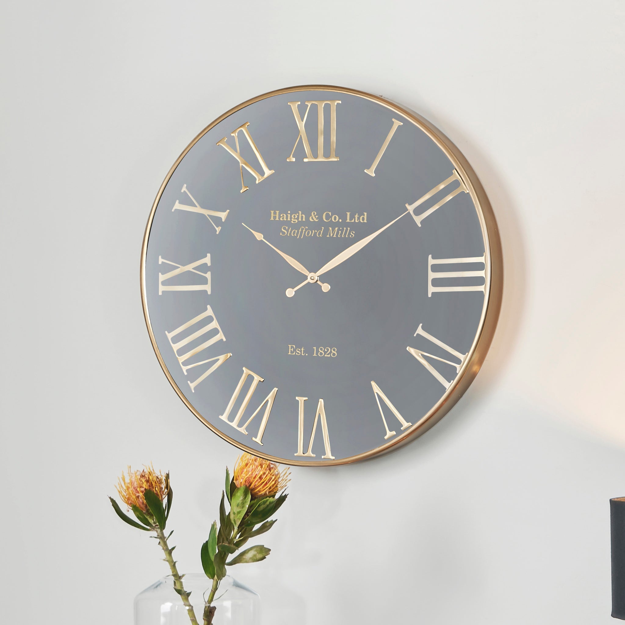 The Antique Gold and Black Metal Wall Clock, featuring a round design with oversized Roman numerals, complements the flowers in a vase below, adding charm to contemporary interiors.