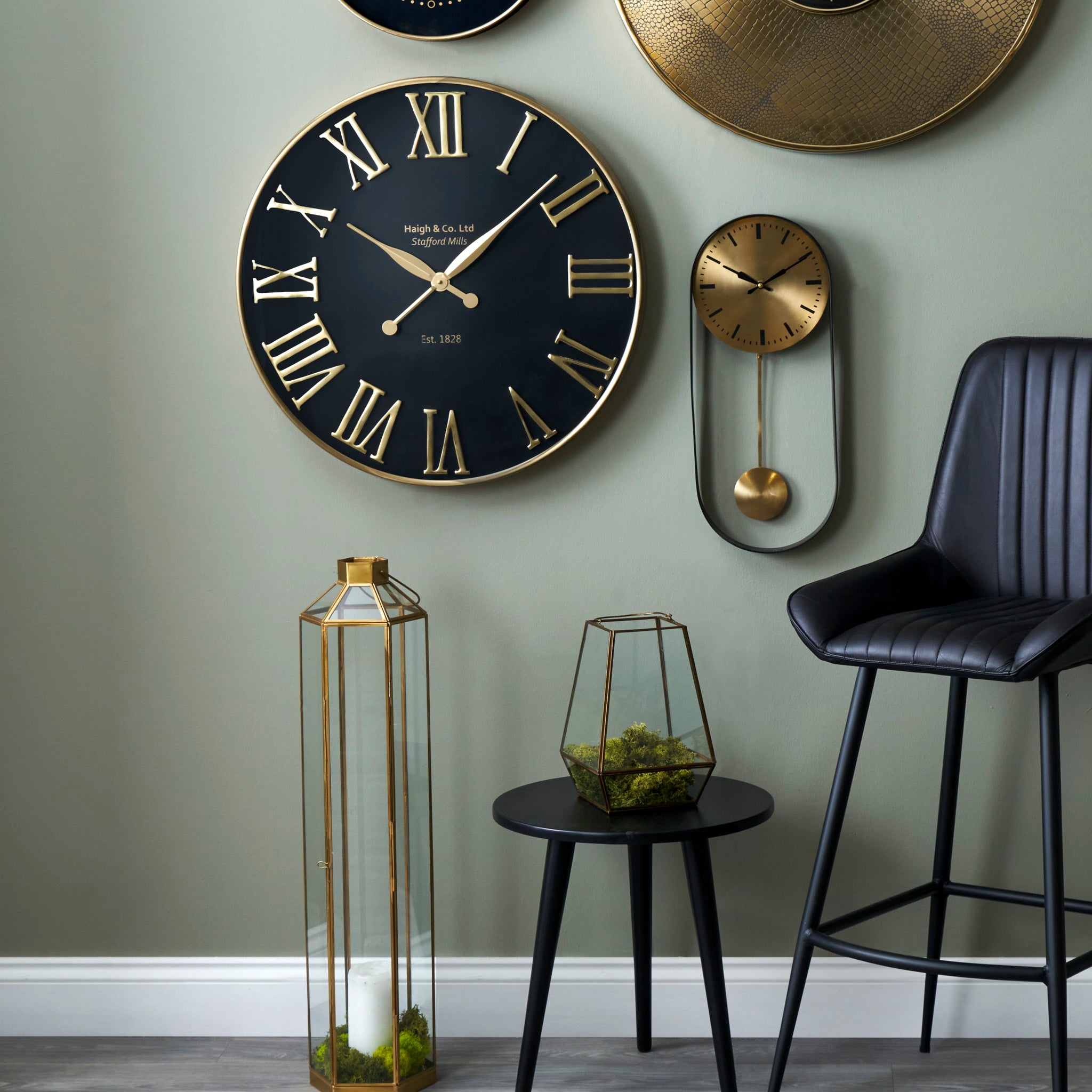 Wall adorned with the Antique Gold and Black Metal Wall Clock featuring oversized Roman numerals, a black chair, a small table, and geometric glass terrariums filled with moss—ideal for contemporary interiors.