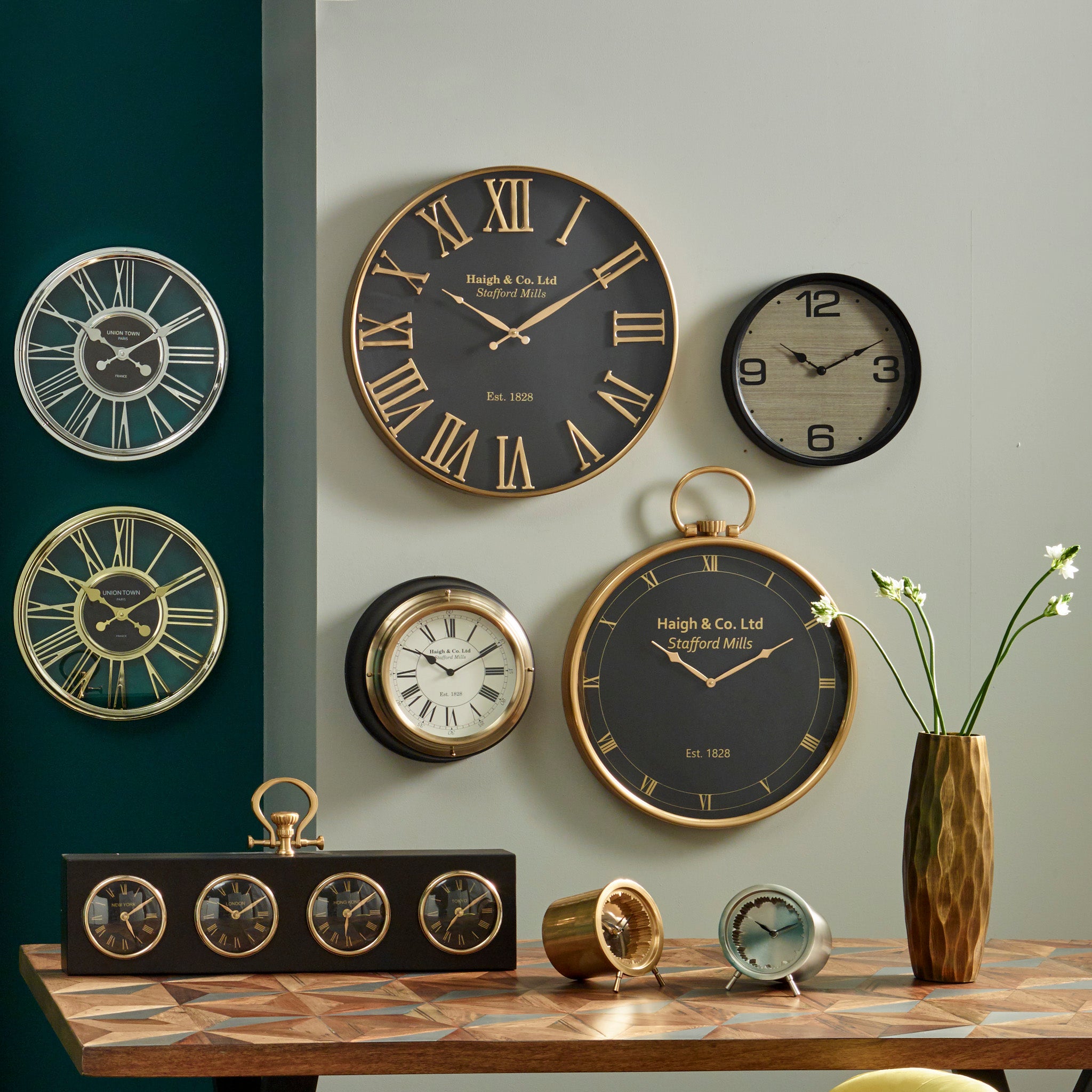 The Antique Gold and Black Metal Wall Clock, showcasing an elegant design with oversized Roman numerals, pairs beautifully with a table decorated with an antique gold clock and a vase of flowers, making it an ideal choice for contemporary interiors.