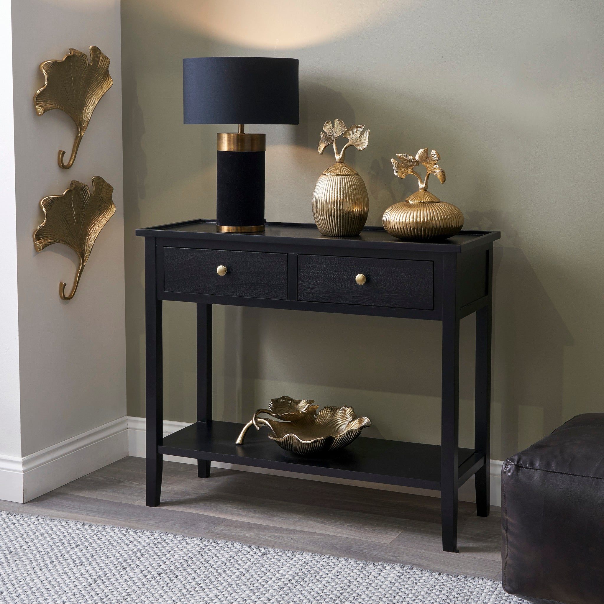 The Fairford Satin Black Pine Wood 2 Drawer Console Table brings elegance with its sleek design, complemented by a stylish black lamp. Mid-century inspired wall-mounted ginkgo leaf hooks enhance the sophistication of the neutral wall backdrop.