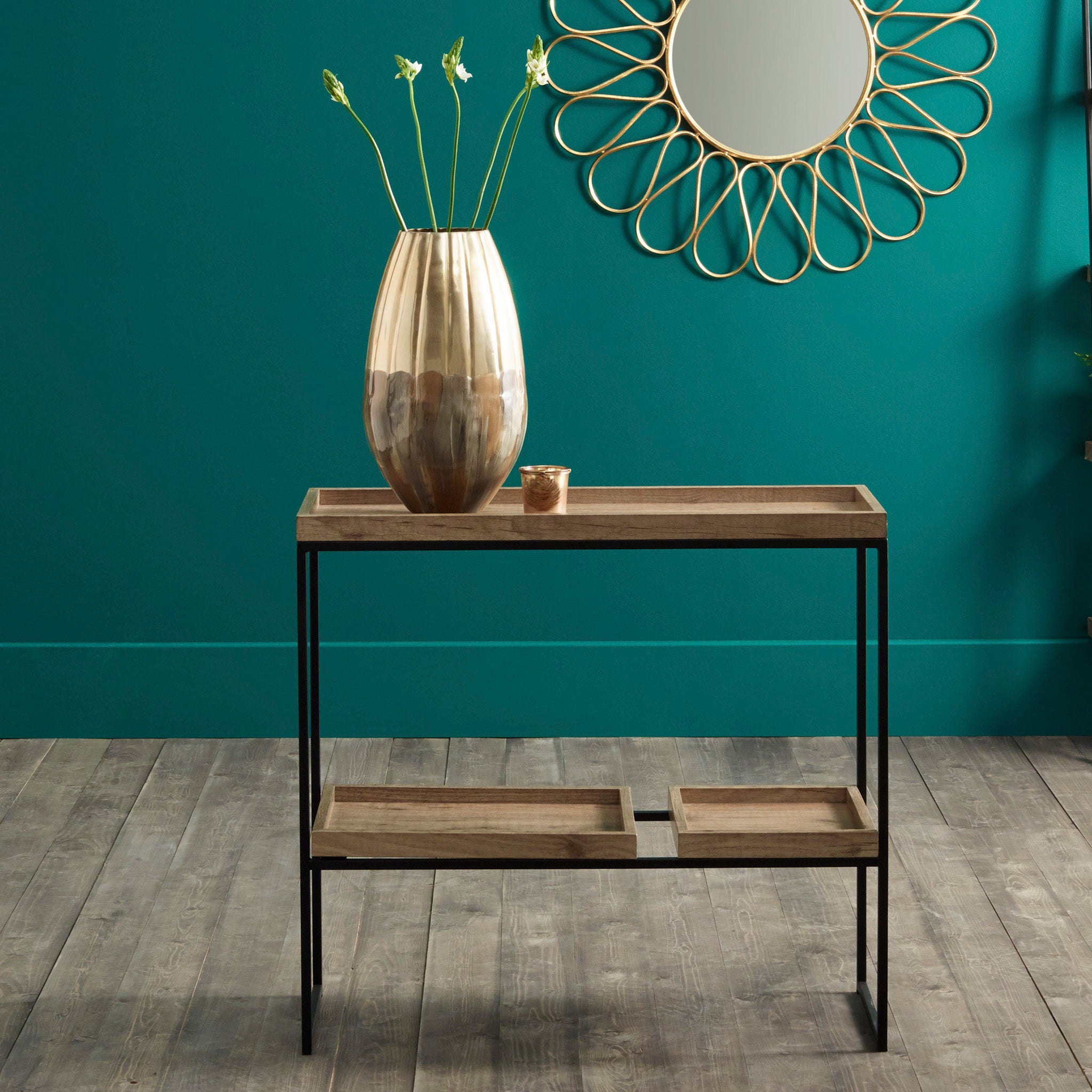 The Pacific Lifestyle Gallery Natural Wood Veneer & Black Metal 2 Shelf Unit, paired with a gold vase of green stems under a decorative mirror on a teal wall, showcases multifunctional design at its finest.