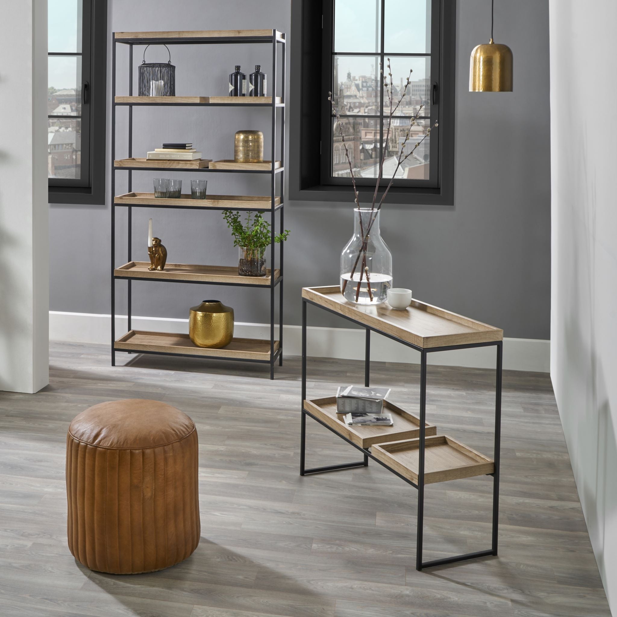 Modern living room with a Scandinavian-styled Gallery Natural Wood Veneer & Black Metal 2 Shelf Unit, a small table with a vase, a brown pouf, and large windows with black frames.