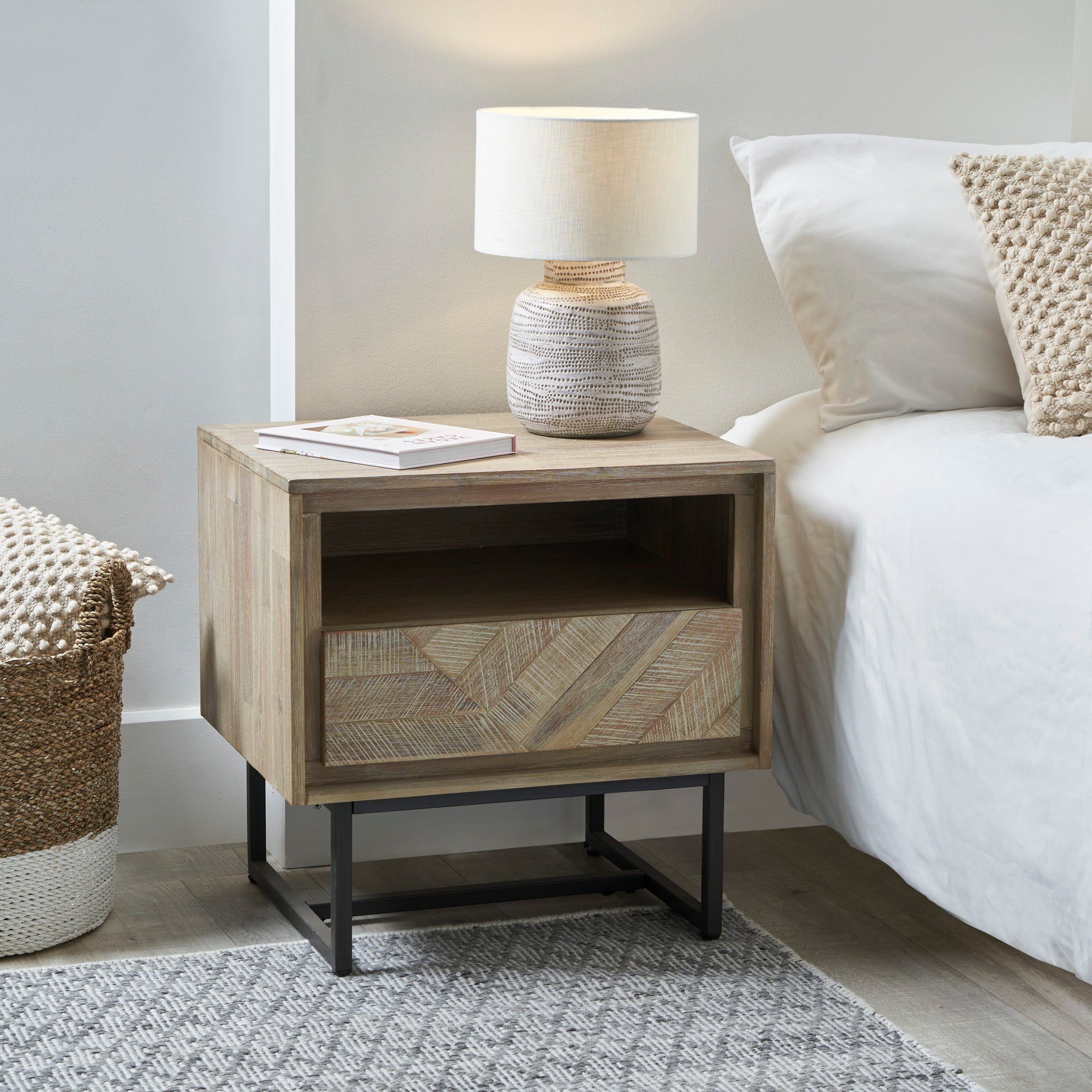 The Marca Sand Wash Acacia Wood and Black Metal Bedside Unit, featuring a geometric drawer and a basket, topped with a white lamp, stands next to a bed adorned with beige pillows, offering ample storage space.