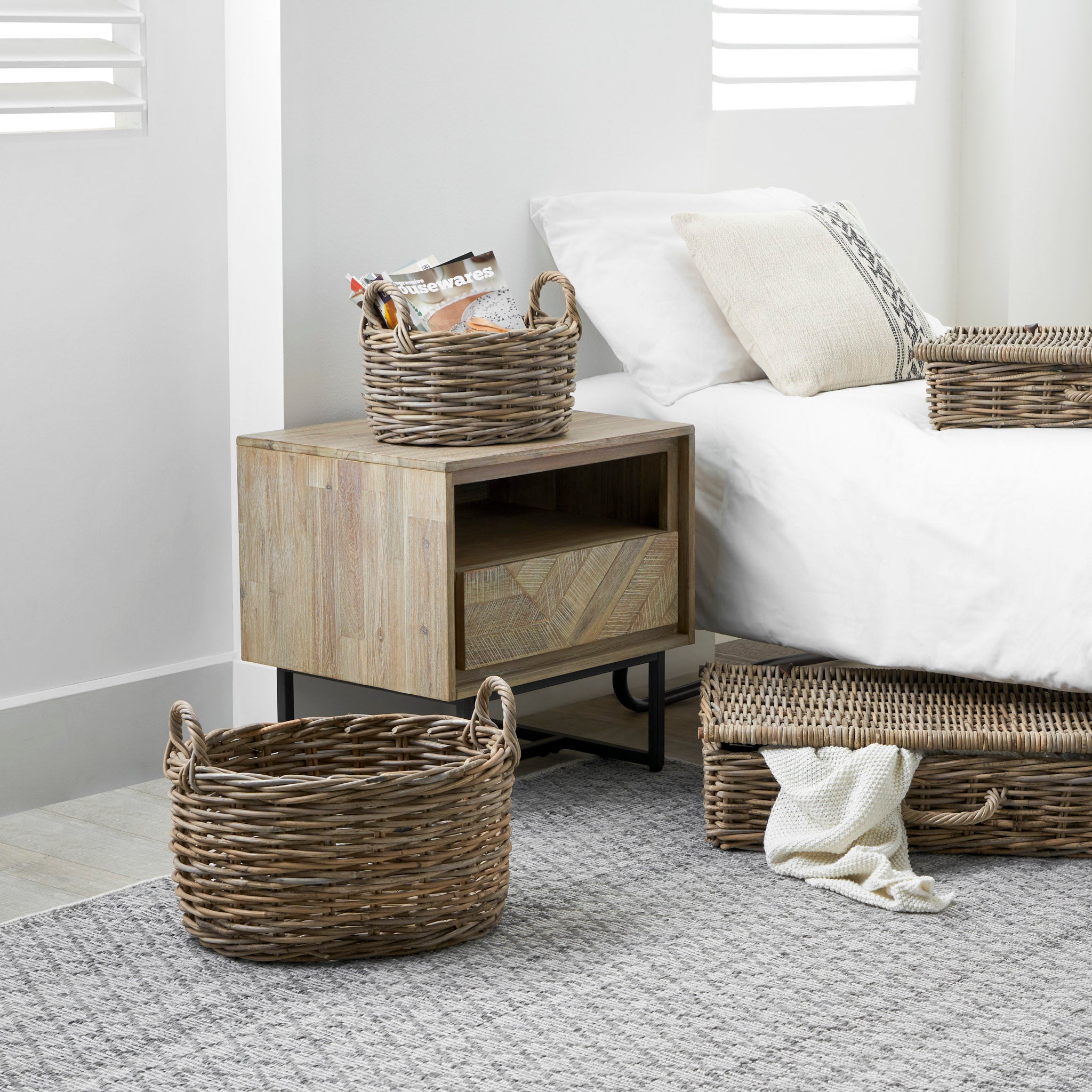 A Scandi-style bedroom featuring a Marca Sand Wash Acacia Wood and Black Metal Bedside Unit, wicker baskets, a bed with white linens, and a knitted blanket on a gray rug offers ample storage space.