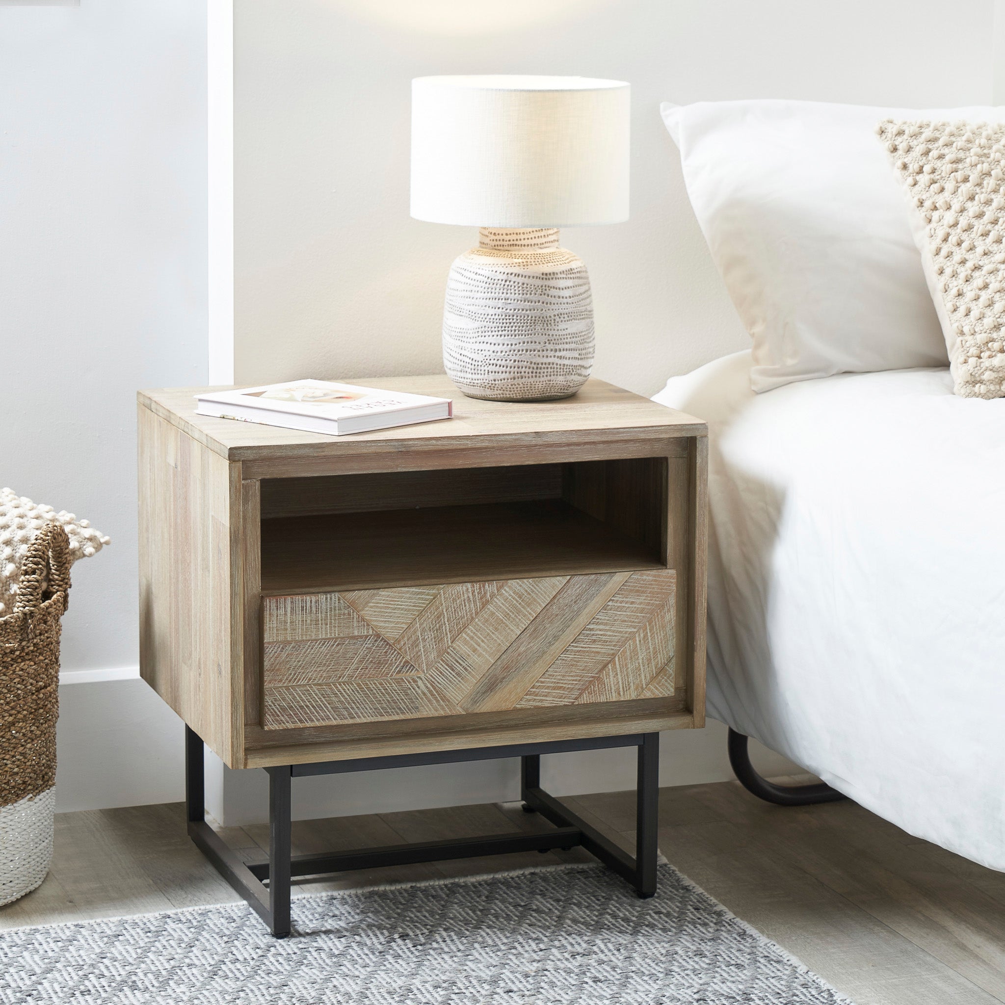 A Marca Sand Wash Acacia Wood and Black Metal Bedside Unit in Scandinavian style features a patterned drawer, ample storage space with an open shelf, a lamp, and a book, situated next to a bed adorned with a textured pillow.