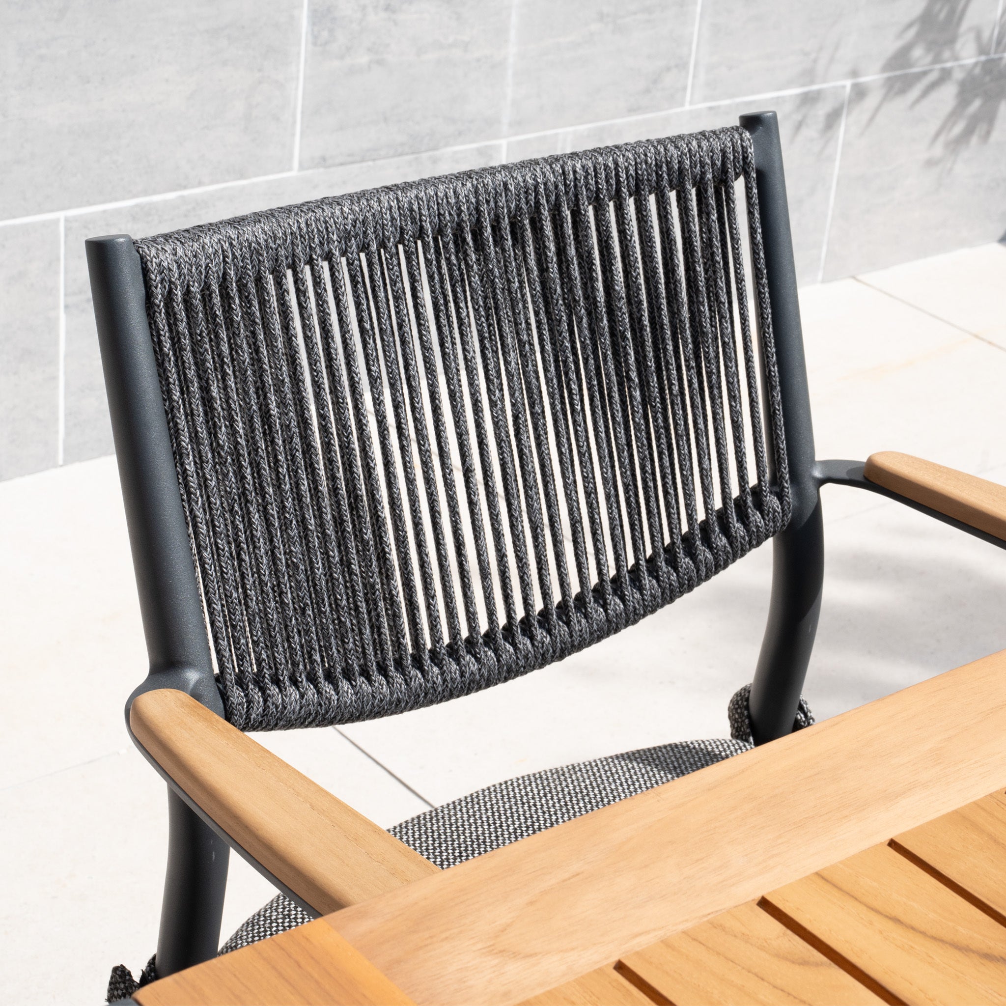 A modern chair with a weather-resistant rope backrest and wooden armrests enhances a wooden table on the tiled patio, ideal for evening relaxation with the Tellaro 8 Seat Rectangular Teak Firepit Dining Set in Charcoal.