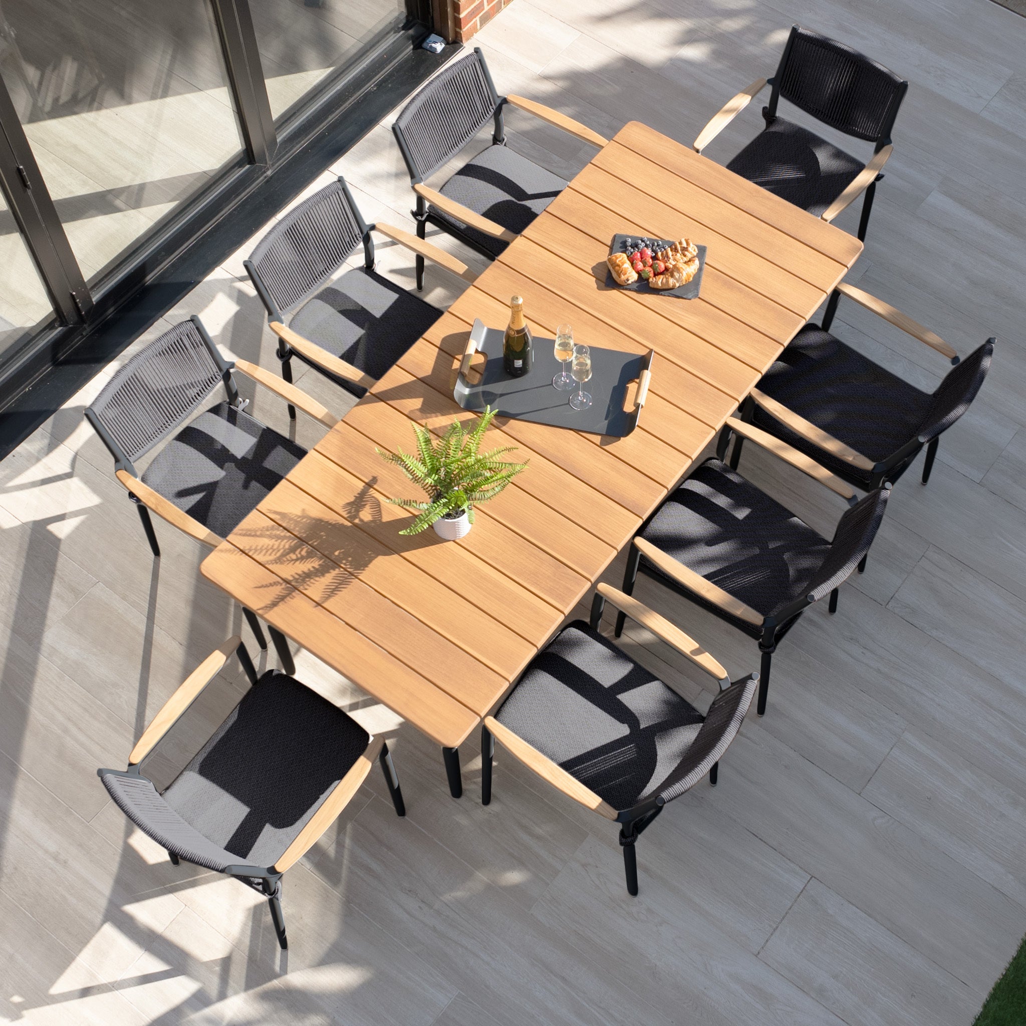 The Tellaro 8 Seat Rectangular Dining Set with Teak Table in Charcoal includes a sleek teak table and eight chairs, complete with wine, snacks, and a potted plant on the patio, ideal for stylish and comfortable outdoor dining.