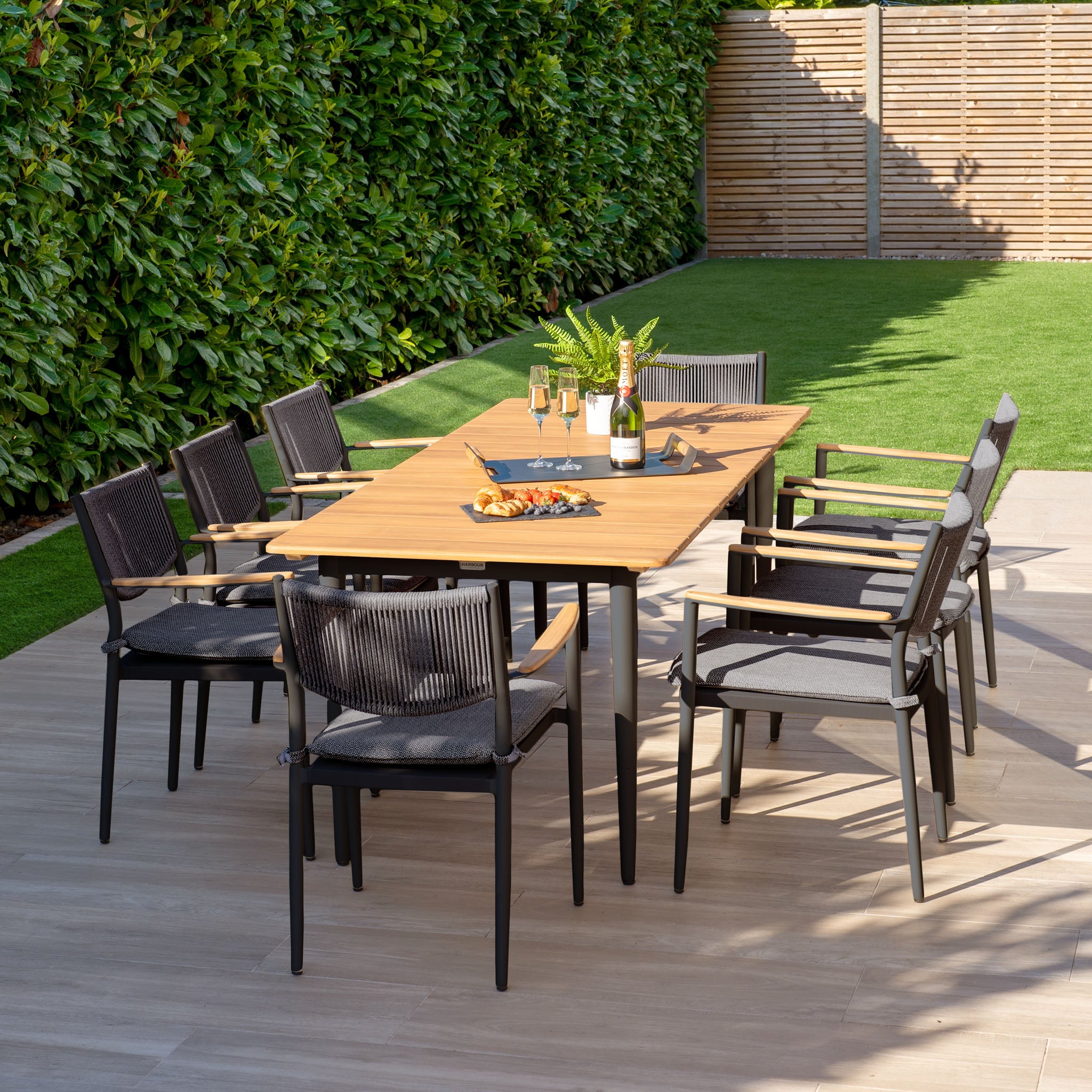 Outdoor dining set featuring the Tellaro 8 Seat Rectangular Dining Set with a weather-resistant teak table in charcoal, surrounded by eight chairs. Champagne and snacks are invitingly arranged on top, amidst lush greenery.