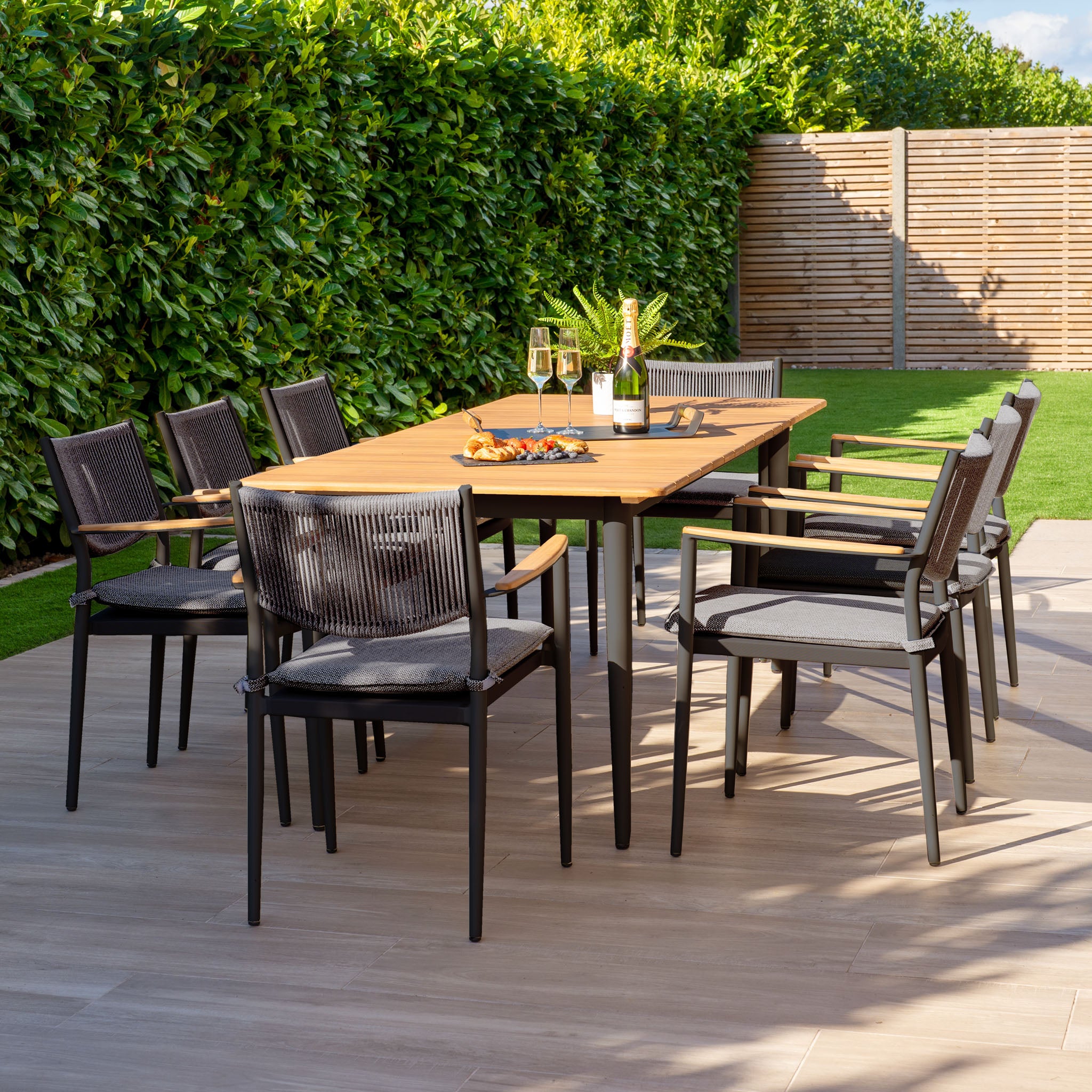 Enjoy outdoor dining with the Tellaro 8 Seat Rectangular Dining Set in Charcoal, featuring a sleek teak table and eight black chairs, wine glasses, and a bottle on a sunny patio.