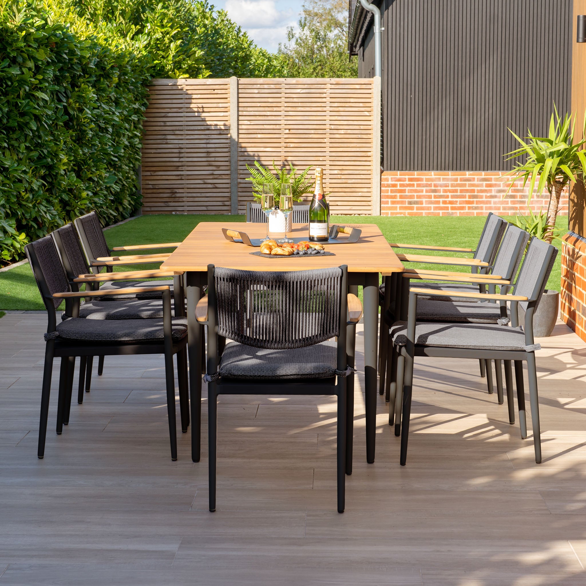 Enjoy refined outdoor dining with the Tellaro 8 Seat Rectangular Dining Set, featuring a stunning teak table in charcoal. This setup, surrounded by lush plants, a brick wall, and a green hedge backdrop, perfectly merges comfort and style for any occasion.