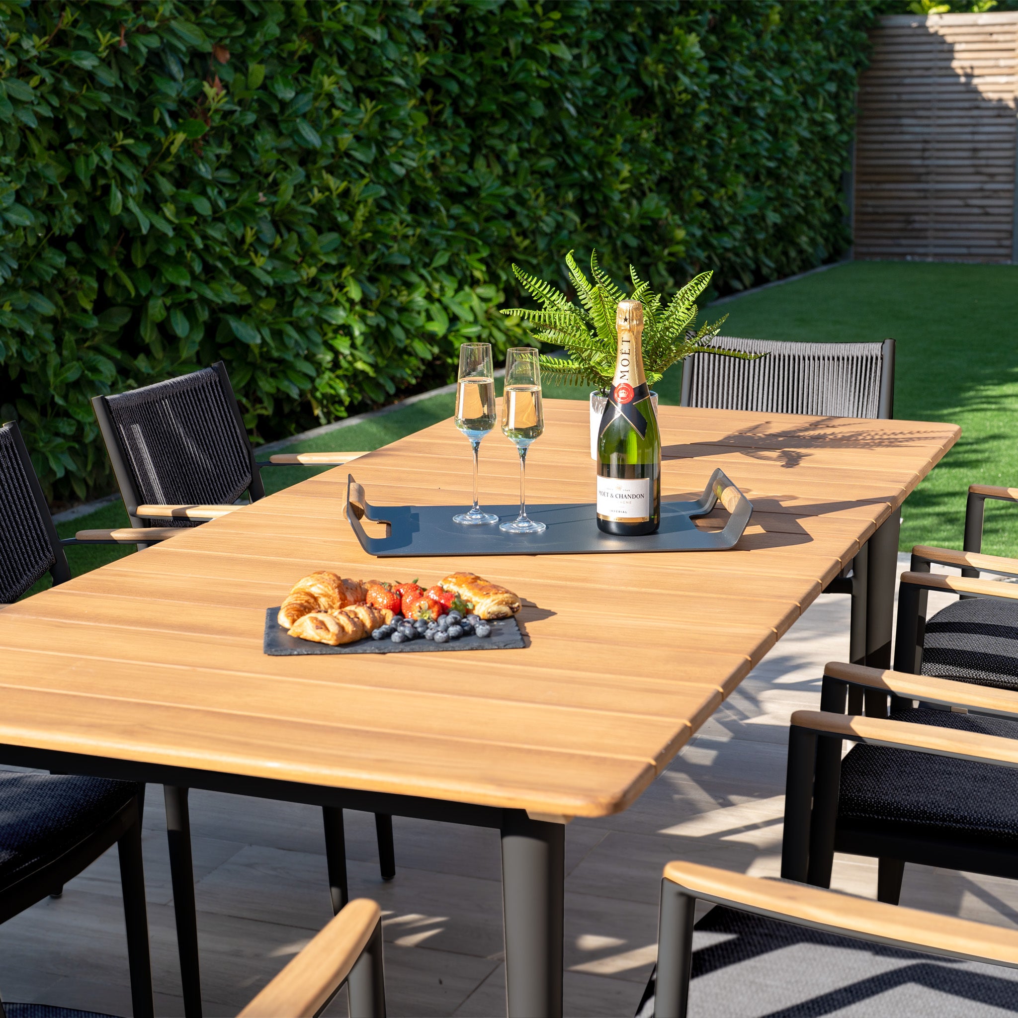 Enhance your outdoor dining with the Tellaro 8 Seat Rectangular Dining Set in Charcoal. A teak table complements croissants, grapes, and champagne, while surrounding chairs invite you to enjoy nature's bounty amid lush greenery.