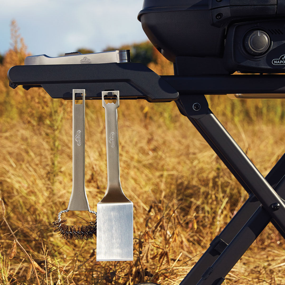 Two grilling tools, a metal brush and a spatula, hang from hooks on the gourmet Napoleon Phantom TravelQ Pro 285X portable gas barbecue in a grassy outdoor setting.