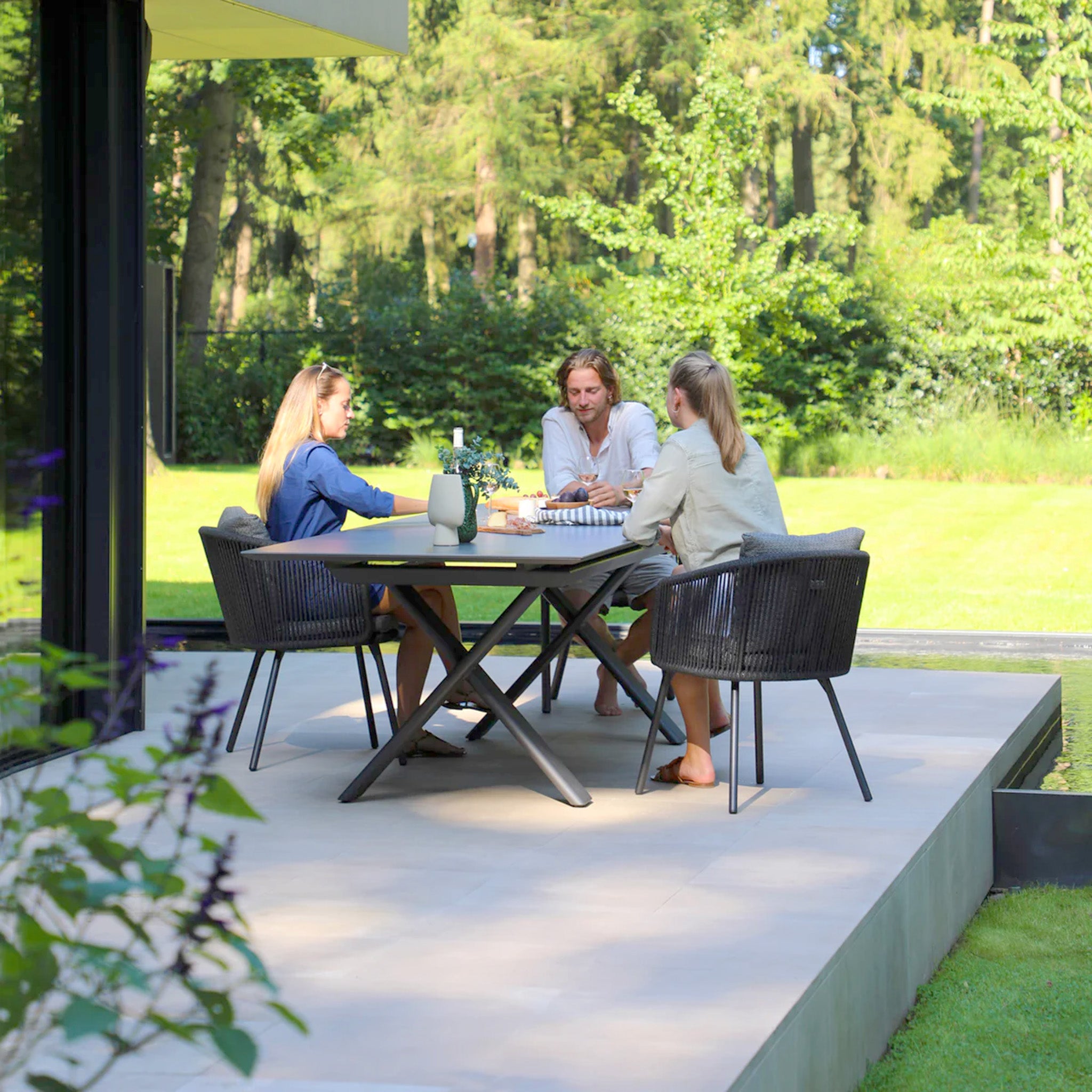 Three people enjoy the serene outdoor space at a patio table, surrounded by greenery and trees. They relax on the Dekota 8 Seat Rope Extending Dining Set in Charcoal, with ergonomically shaped chairs complementing the scenic beauty.