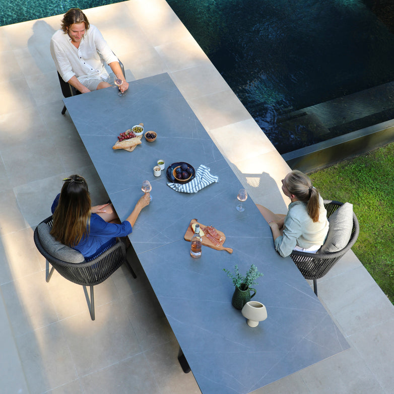 Three people are seated around a long outdoor table with snacks and drinks beside a pool, enjoying the garden ambiance. The setting features the Dekota 8 Seat Rope Extending Dining Set in Charcoal, complete with ergonomically shaped chairs for added comfort.