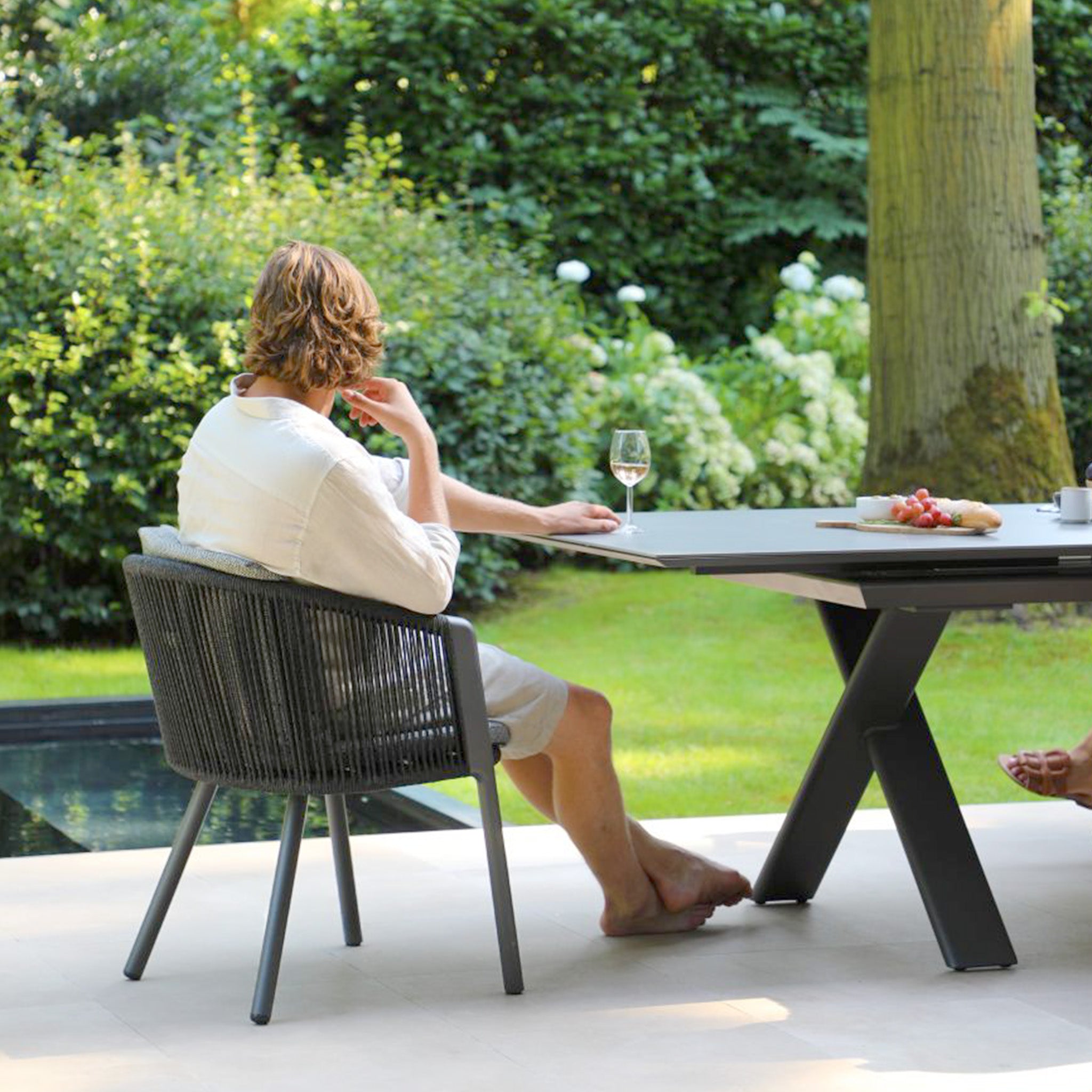 A person relaxes in an ergonomic chair from the Dekota 8 Seat Rope Extending Dining Set in Charcoal at an outdoor table with wine, gazing at a grassy area surrounded by lush greenery.