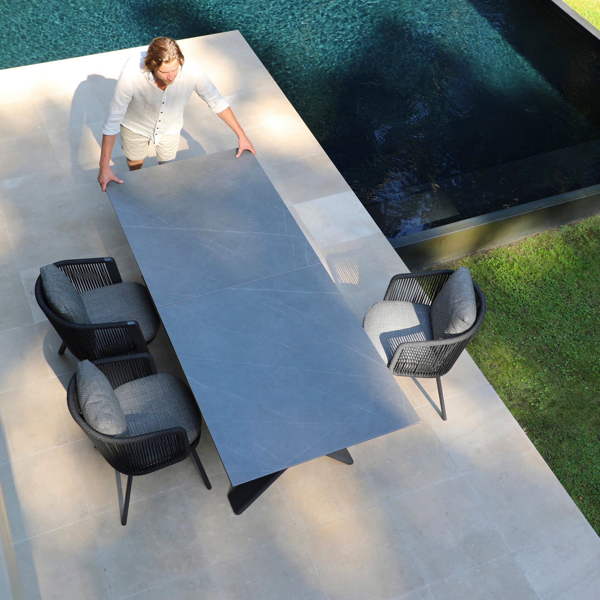 A person arranges a sleek, charcoal outdoor table from the Dekota 8 Seat Rope Extending Dining Set with three ergonomic chairs on a patio near the pool, enhancing the inviting outdoor space.