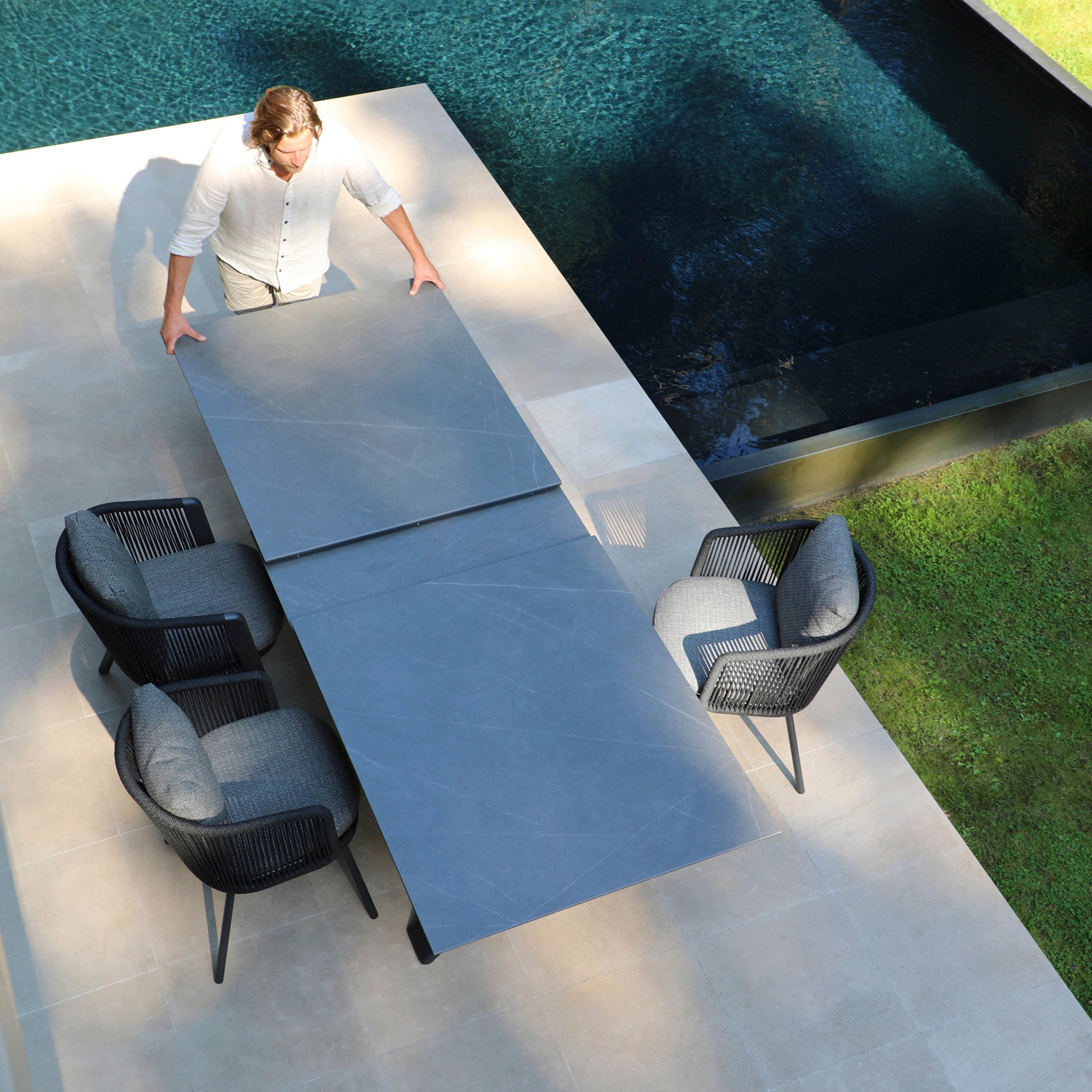 A man adjusts a Dekota 8 Seat Rope Extending Dining Set in Charcoal on a patio next to a pool, featuring eight ergonomically designed chairs for optimal comfort in your outdoor space.