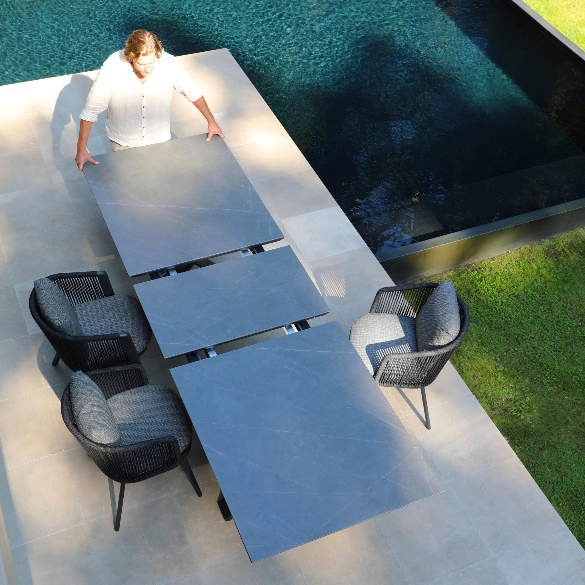 A person sets up a Dekota 8 Seat Rope Extending Dining Set in Charcoal, featuring four ergonomically shaped chairs, near their pool.