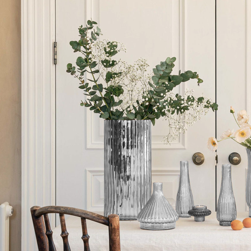 A large silver vase with flowers sits on a table, flanked by several smaller Daphne Ribbed Vases in Clear, against a white door backdrop. This 1960s inspired design adds a touch of nostalgia and elegance to any room while embracing sustainable décor principles.