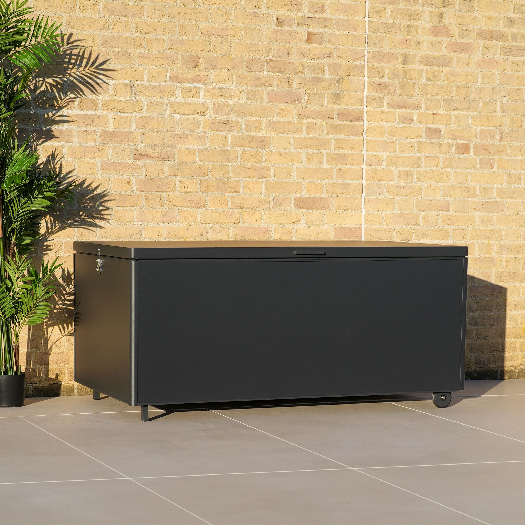 A large grey outdoor storage box with aluminium construction sits against a brick wall, accompanied by a potted plant on the left side.