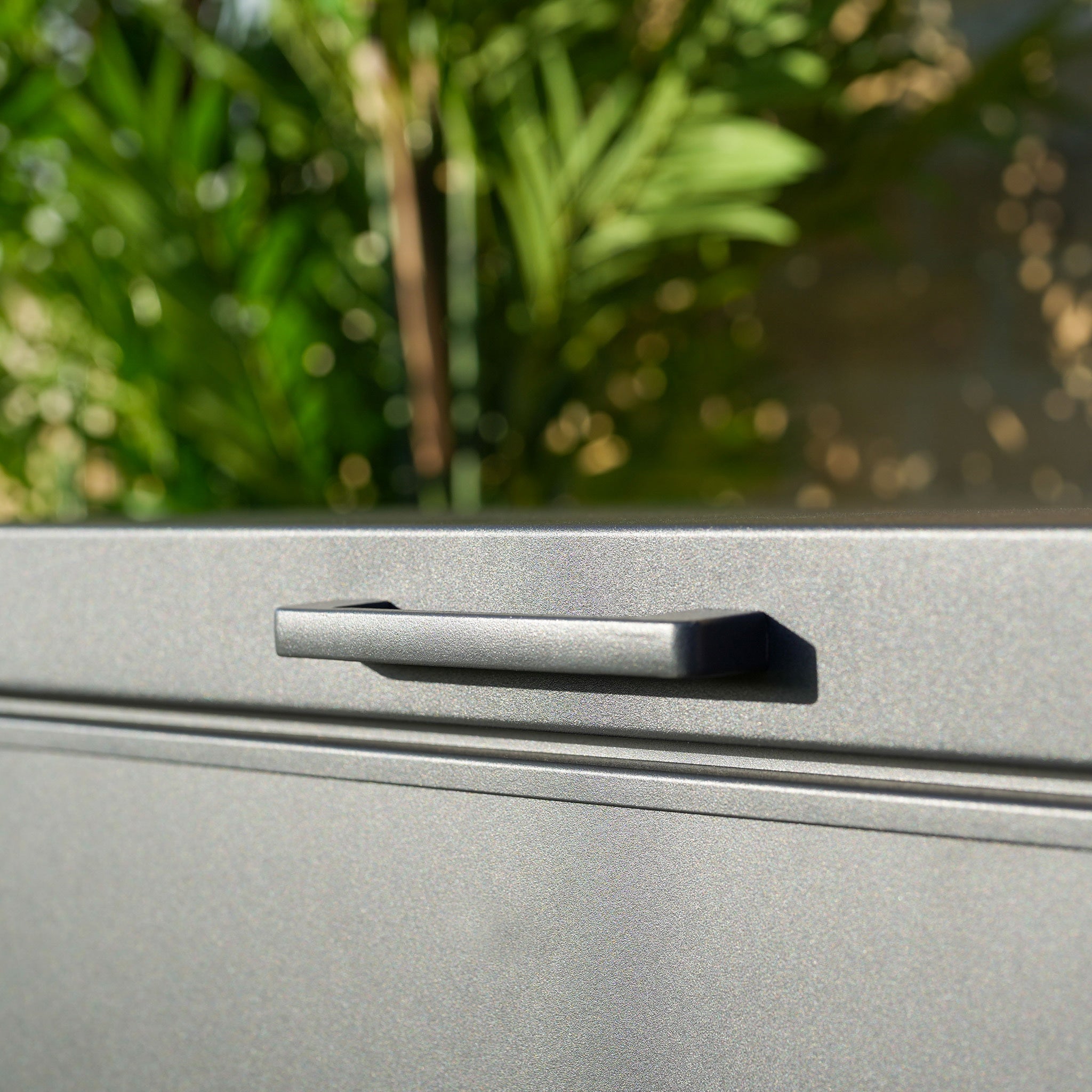 Close-up of a sleek black drawer handle on a grey Large Aluminium Storage Box with blurred green foliage in the background, highlighting its sturdy aluminium construction.