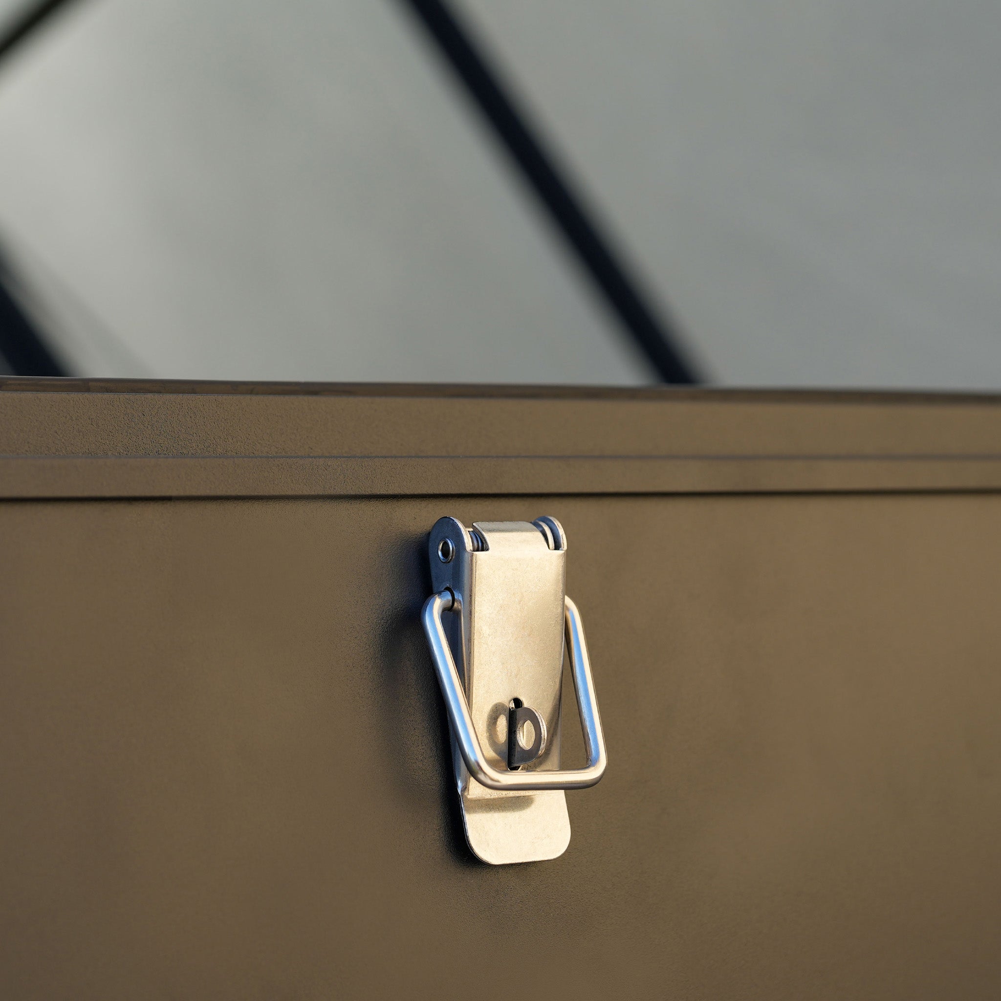 Close-up of a metal clasp latch on the Large Aluminium Storage Box in Grey, set against a blurred geometric background, highlighting its durable aluminium construction.