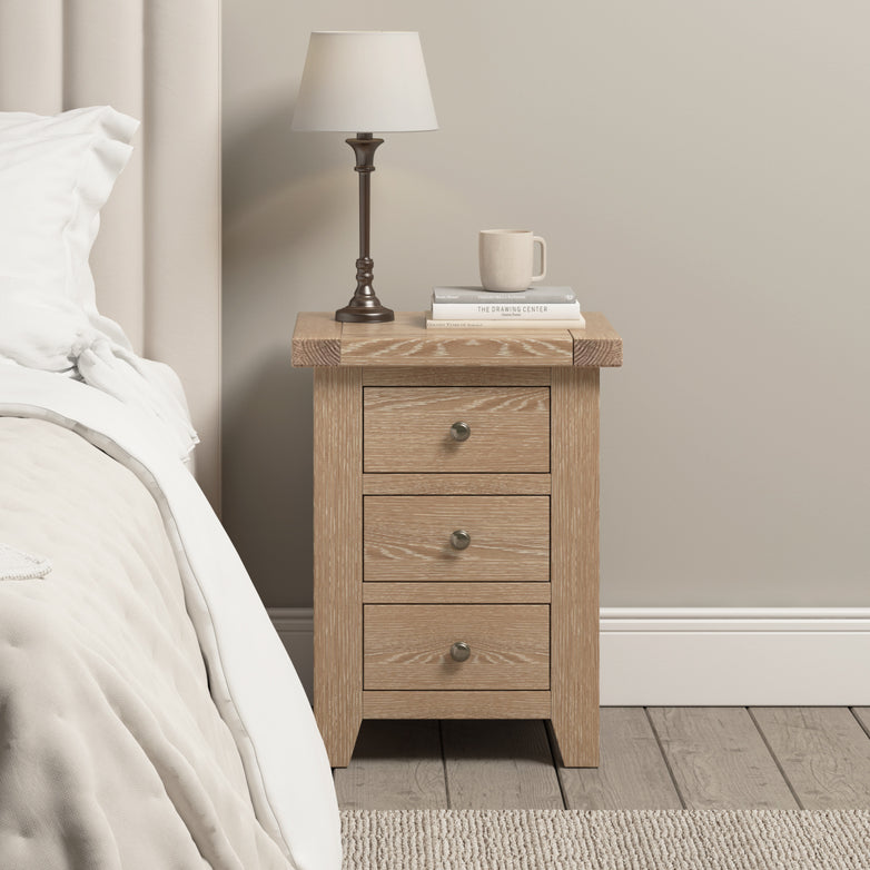 Introducing the Burford 3 Drawers Bedside Table in Natural Oak. This stylish piece, featuring a whitewash finish, is perfect beside your bed and provides ample storage with its three spacious drawers. Adorned with a lamp, mug, and books, it adds both functionality and charm to any room’s wooden floor.