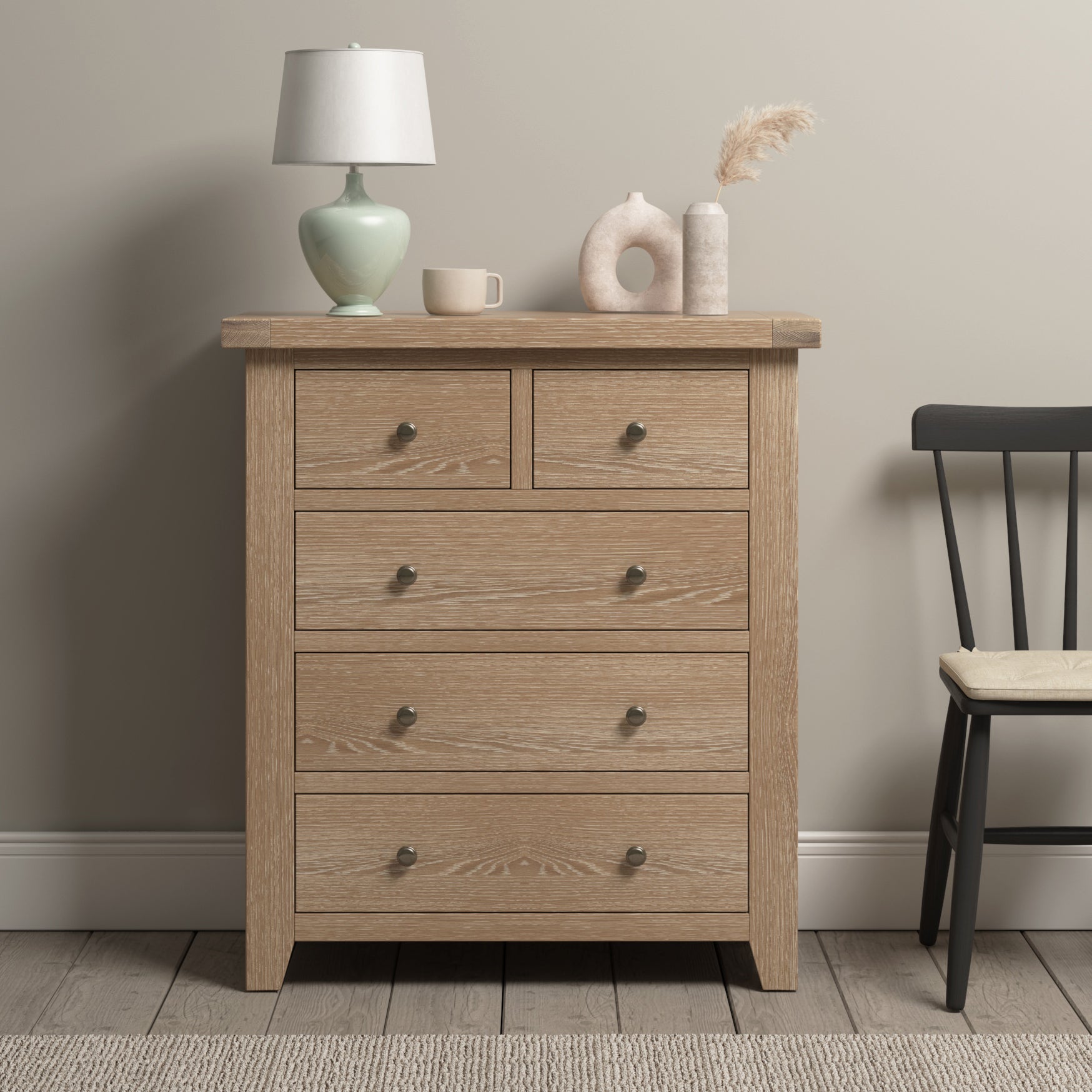 A Burford 2 Over 3 Chest of Drawers in Natural Oak with five drawers, a table lamp, a ceramic sculpture, and a vase stands next to a black chair with a cushion. This chest of drawers offers ample storage solutions for any room.