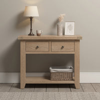 A Burford 2 Drawers Console Table in Natural Oak, featuring two storage drawers, a basket on the bottom shelf, a lamp, a cup, dried flowers, and a framed picture.