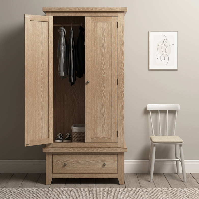 The Burford Double Wardrobe with Drawer in Natural Oak, featuring open doors that reveal hanging clothes and a basket inside, stands adjacent to a white chair and wall art. This piece of solid oak furniture infuses charm and functionality into any room.