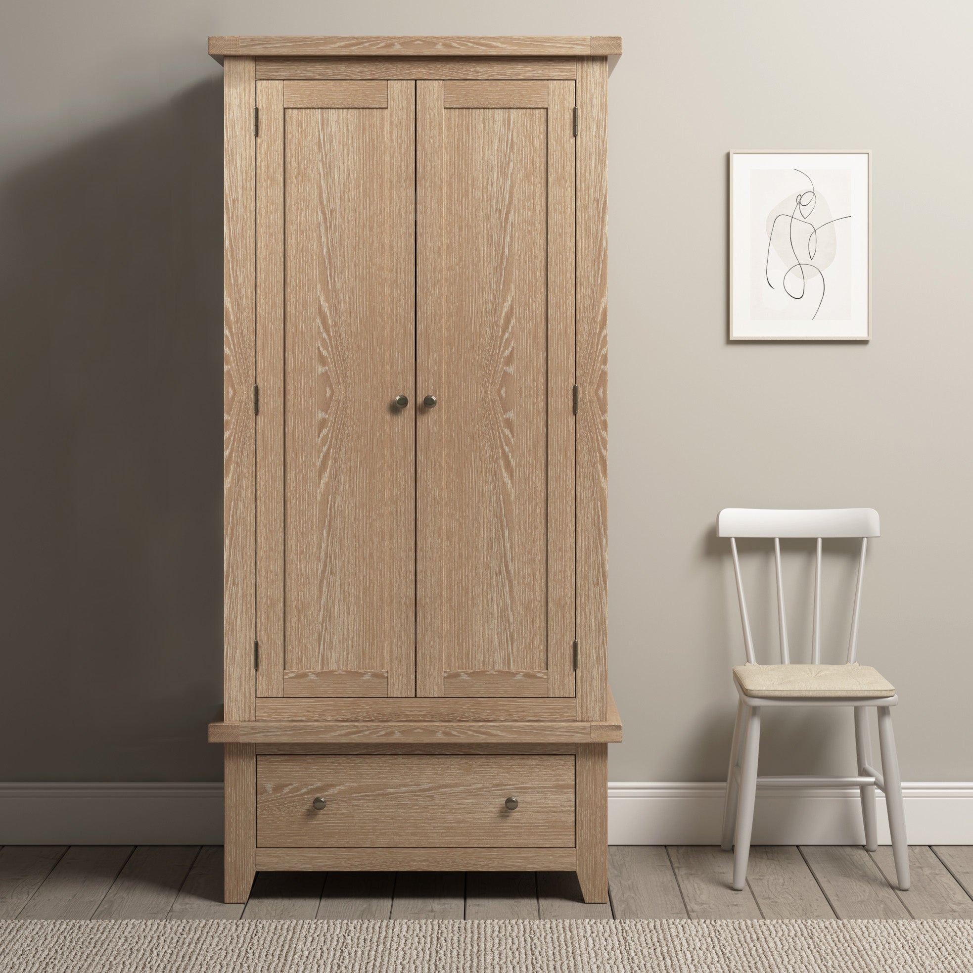 The Burford Double Wardrobe with Drawer in Natural Oak stands next to a white chair against a beige wall adorned with framed art.