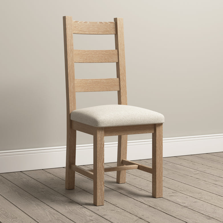 The Burford Ladderback Dining Chairs with Fabric Seat in Natural Oak are elegantly placed on a wooden floor against a plain wall, their natural oak finish enhancing the simple yet refined aesthetic of the room.