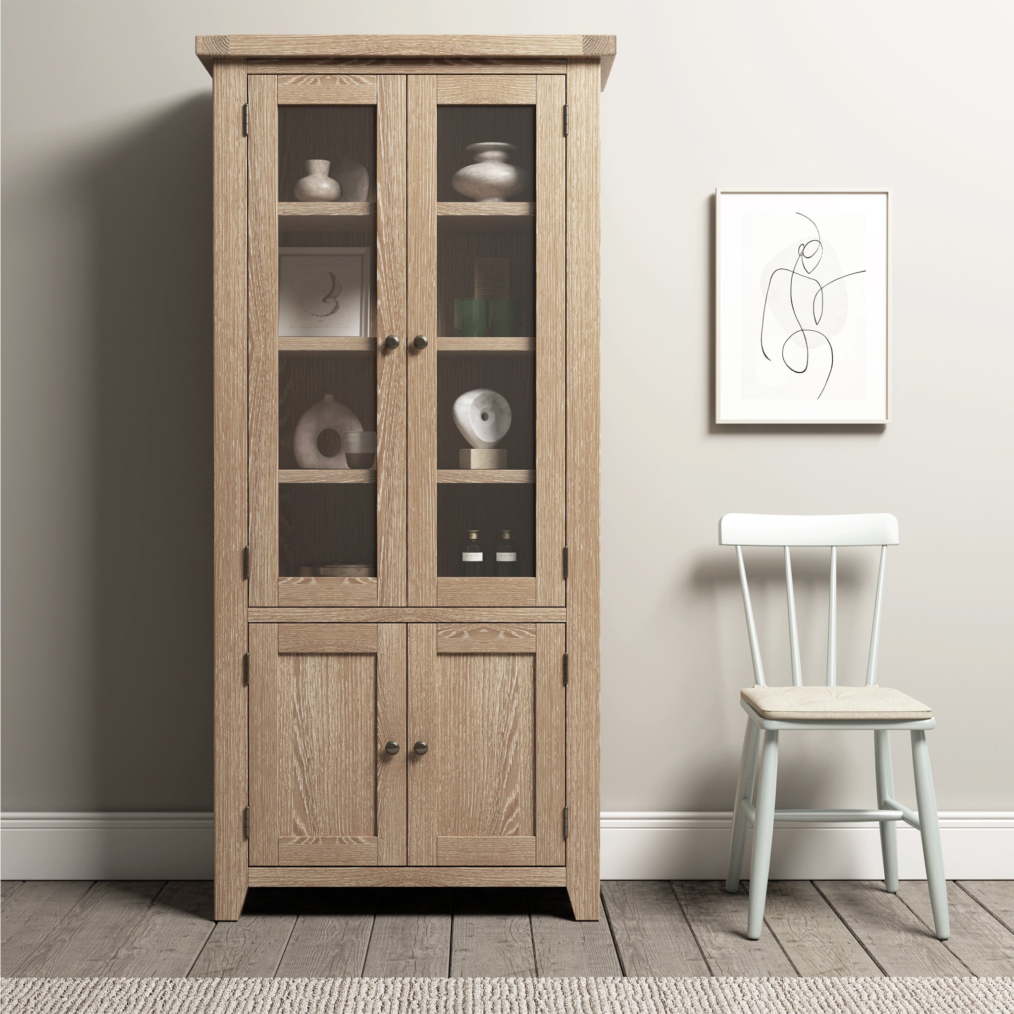 The Burford Display Cabinet in Natural Oak, featuring glass-panelled doors, elegantly displays decor items against a light wall adorned with minimalist framed art, paired with a white chair.