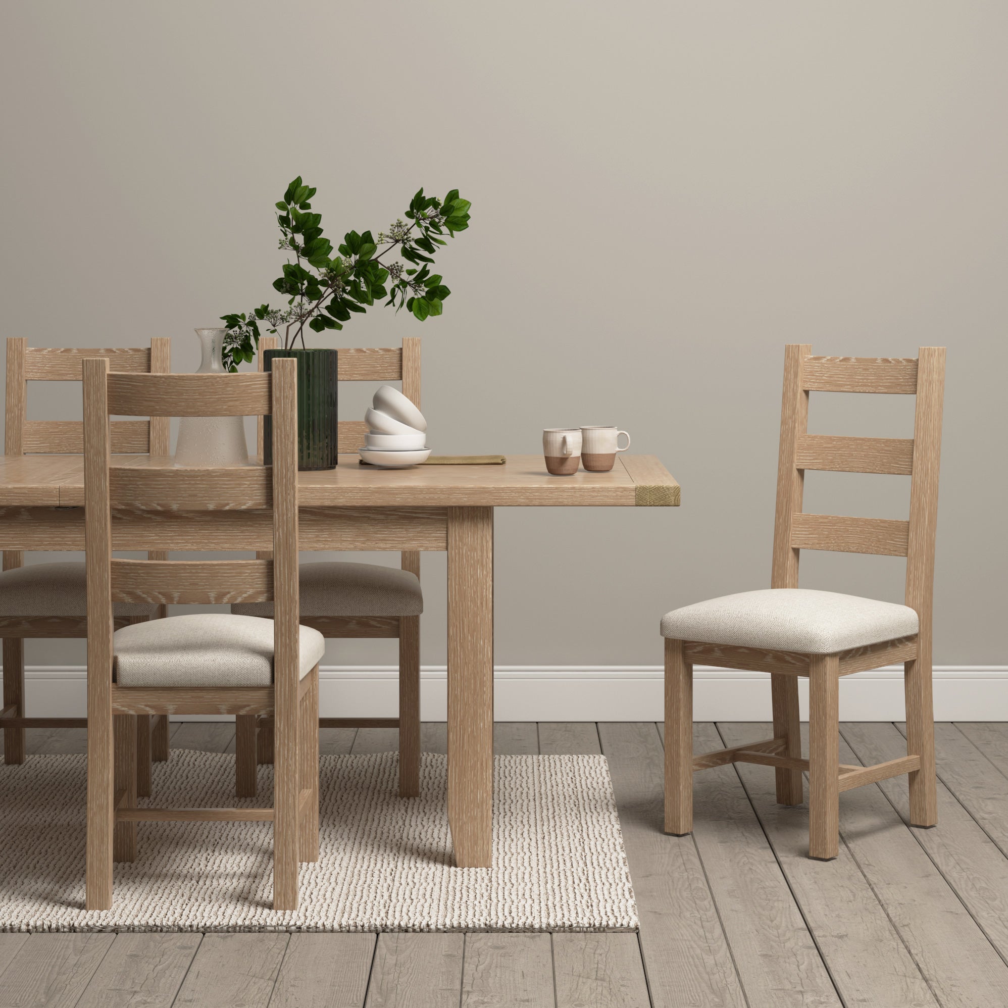 Minimalist dining space featuring a Burford 6-10 Seater Extending Dining Table in Natural Oak with wooden chairs, white rug, green plant in vase, and neutral-colored cups.