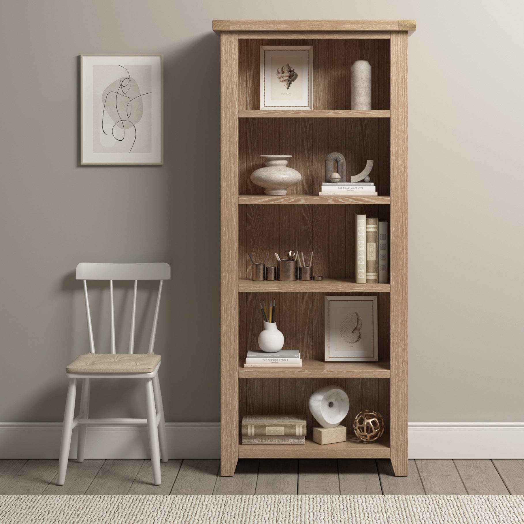 The Burford Large Bookcase in Natural Oak, adorned with art, ceramics, books, and decor, stands beside a white chair with a cushion against a beige wall, embodying contemporary elegance.