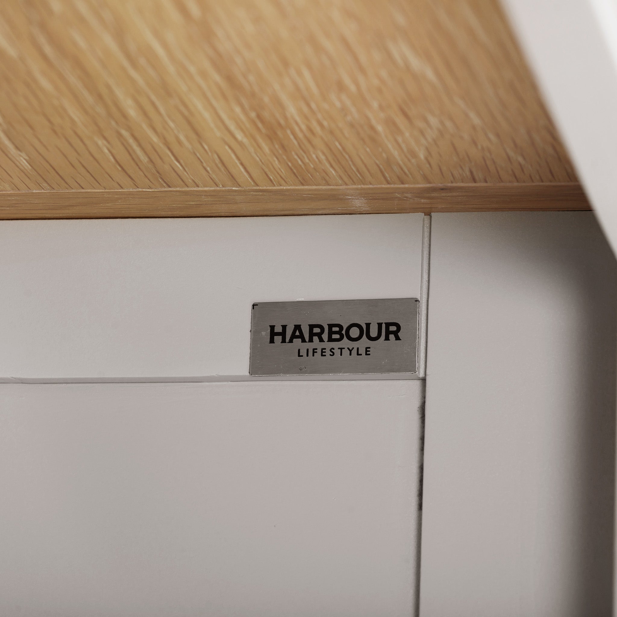 Close-up of a "Harbour Lifestyle" label on a piece of white and wooden furniture, showcasing the elegance of natural oak. The design echoes the sophistication akin to the Burford Large Bookcase in Pebble Grey, blending functional shelving space with timeless style.