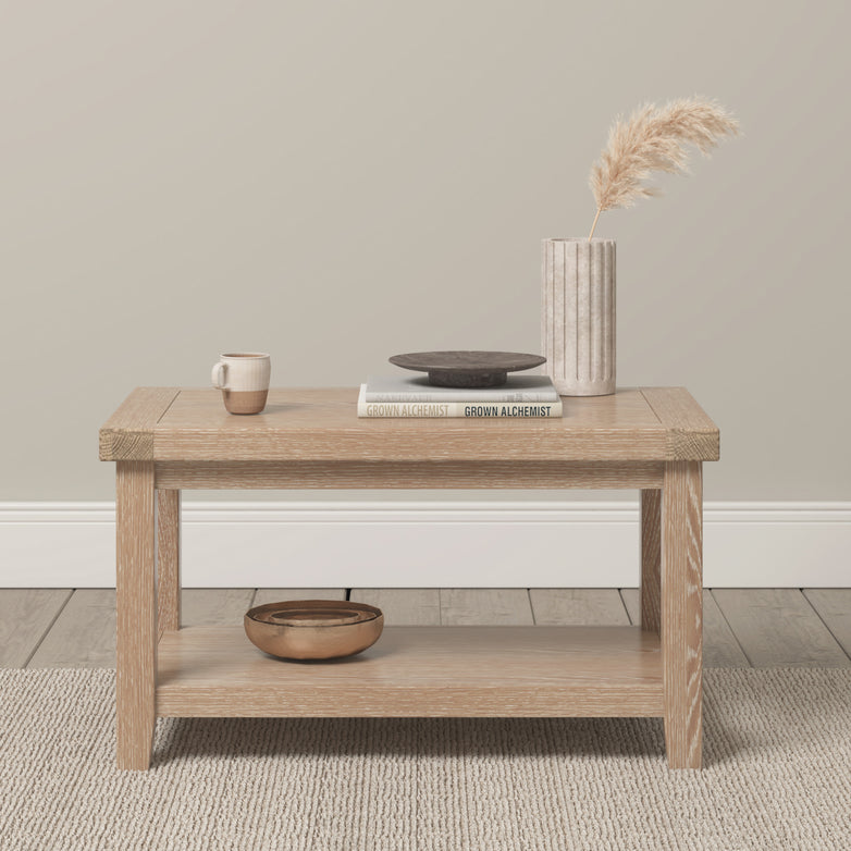 The Burford Large Coffee Table with Shelf in Natural Oak features a minimalist design, adorned with a vase, book, cup, and plate on top. A wooden bowl rests on the lower shelf, all set within a neutral room.