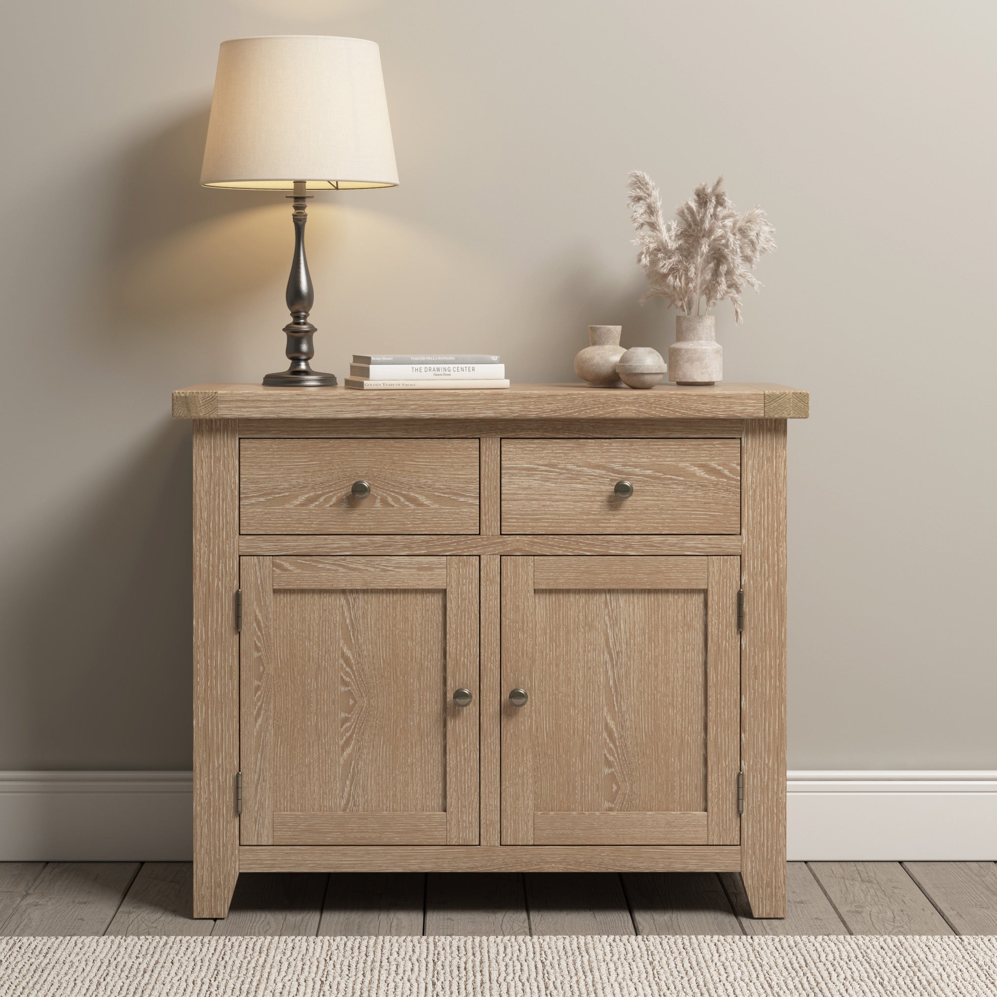 The Burford 2 Door 2 Drawers Sideboard in Natural Oak is a stunning piece featuring two drawers and two cabinet doors. Adorned with a lamp, books, and dried flowers in vases on top, it adds a touch of rustic elegance to any room.