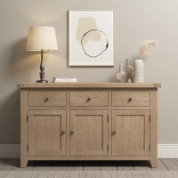 The Burford Large 3 Door 3 Drawers Sideboard in Natural Oak, embellished with a lamp, books, and decorative objects on top and paired with abstract art on the wall above, provides ample storage with its spacious drawers.