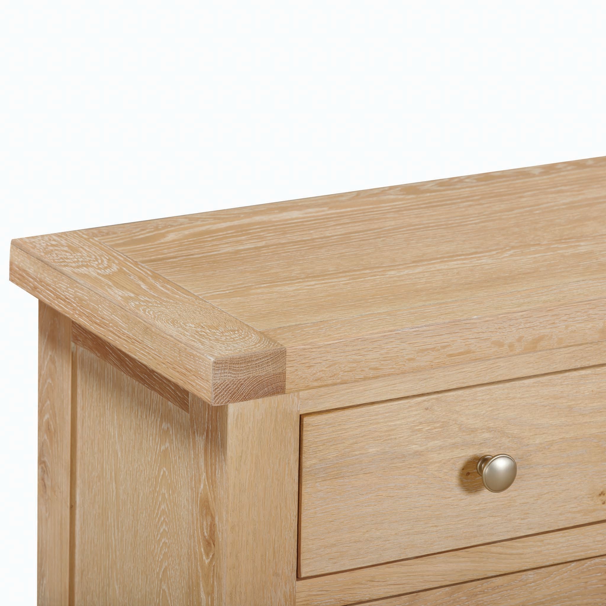 Close-up of the Natural Oak Burford Large 4 Door 3 Drawer Sideboard, showcasing its light wooden finish and simple design. The image highlights a corner and a drawer adorned with a round metal knob, exuding the timeless charm reminiscent of elegant craftsmanship.