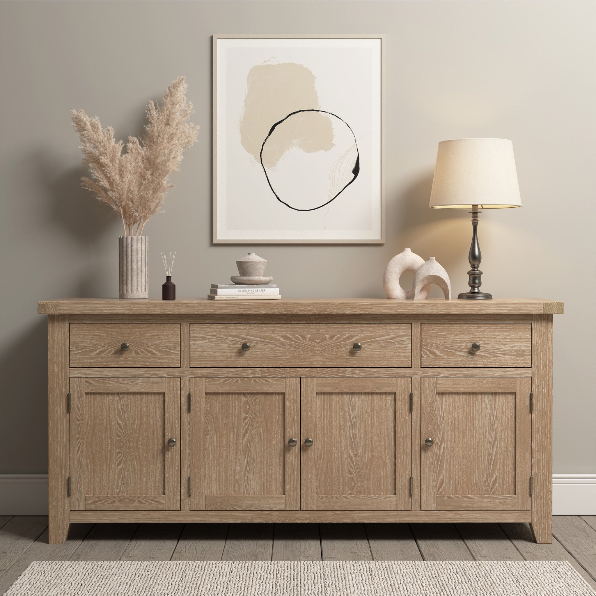 The Burford Large 4 Door 3 Drawers Sideboard in Natural Oak offers versatile storage with its three drawers and four cabinets. Adorning this neutral-toned piece are a stylish lamp, elegant art, and charming decor, all perfectly complemented by a rug beneath.